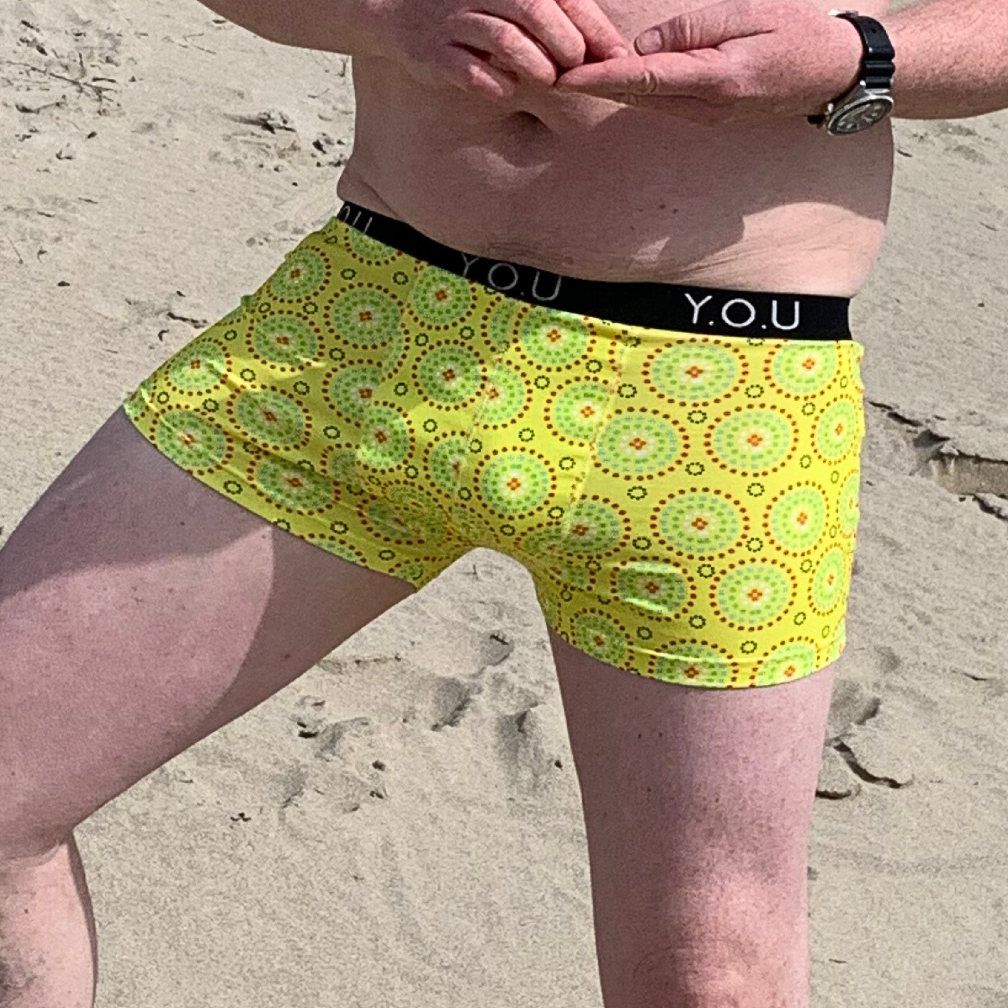 A person standing on a sandy beach wearing yellow men’s hipster trunks with a bright circular pattern. The waistband of the trunks has "Y.O.U" printed on it. The individual is also wearing a wristwatch on their left arm.
