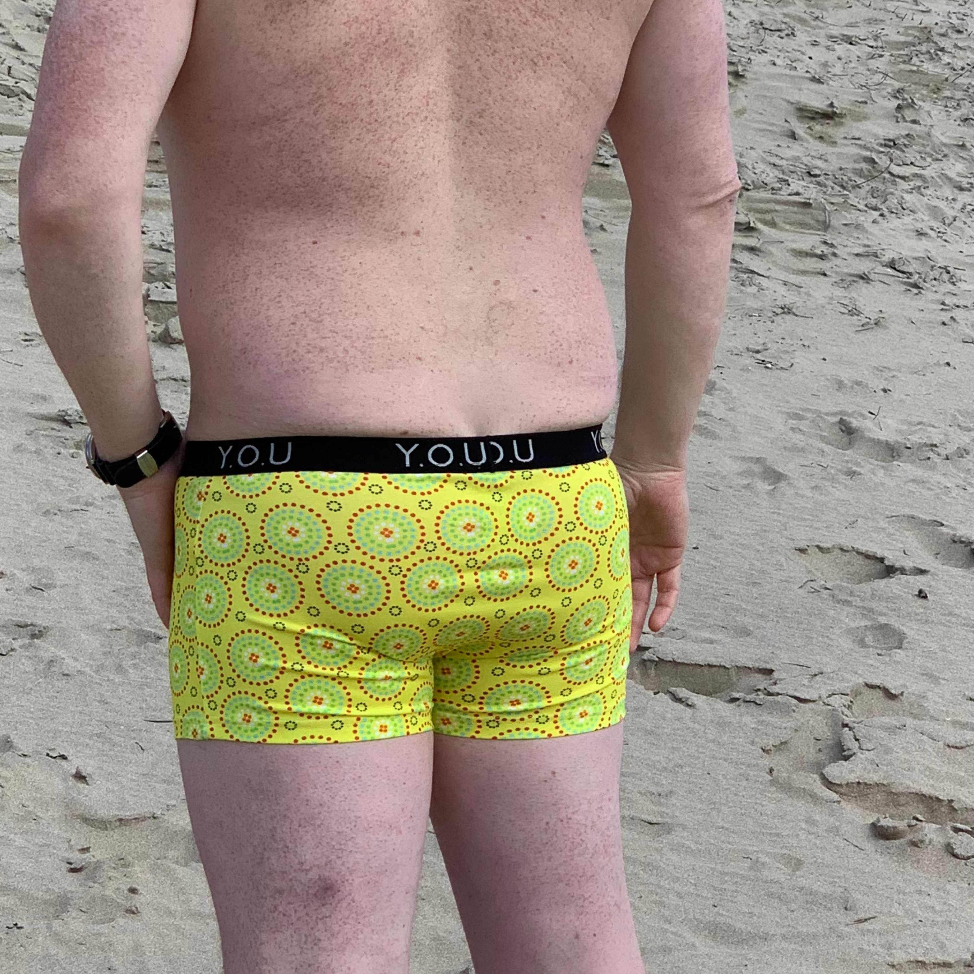 A person with fair skin and freckles stands on a sandy beach. They are facing away from the camera and are wearing bright yellow men's hipster trunks, featuring a colorful pattern that includes red and green circular designs.