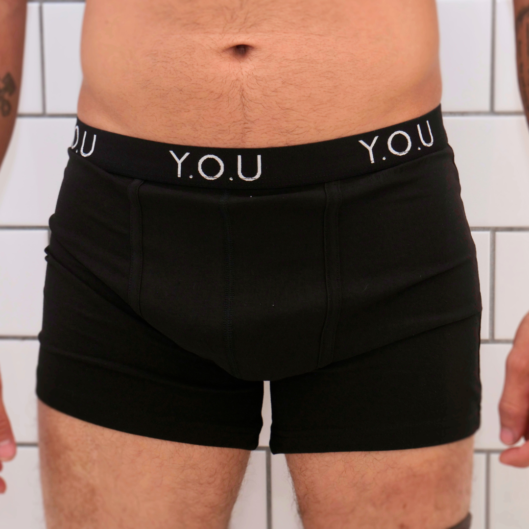 A person is wearing black mid-length trunks with the brand name "Y.O.U" printed on the waistband. The image focuses on the midsection, showcasing two visible tattoos on the left and right arms. The background consists of white tiled walls.
