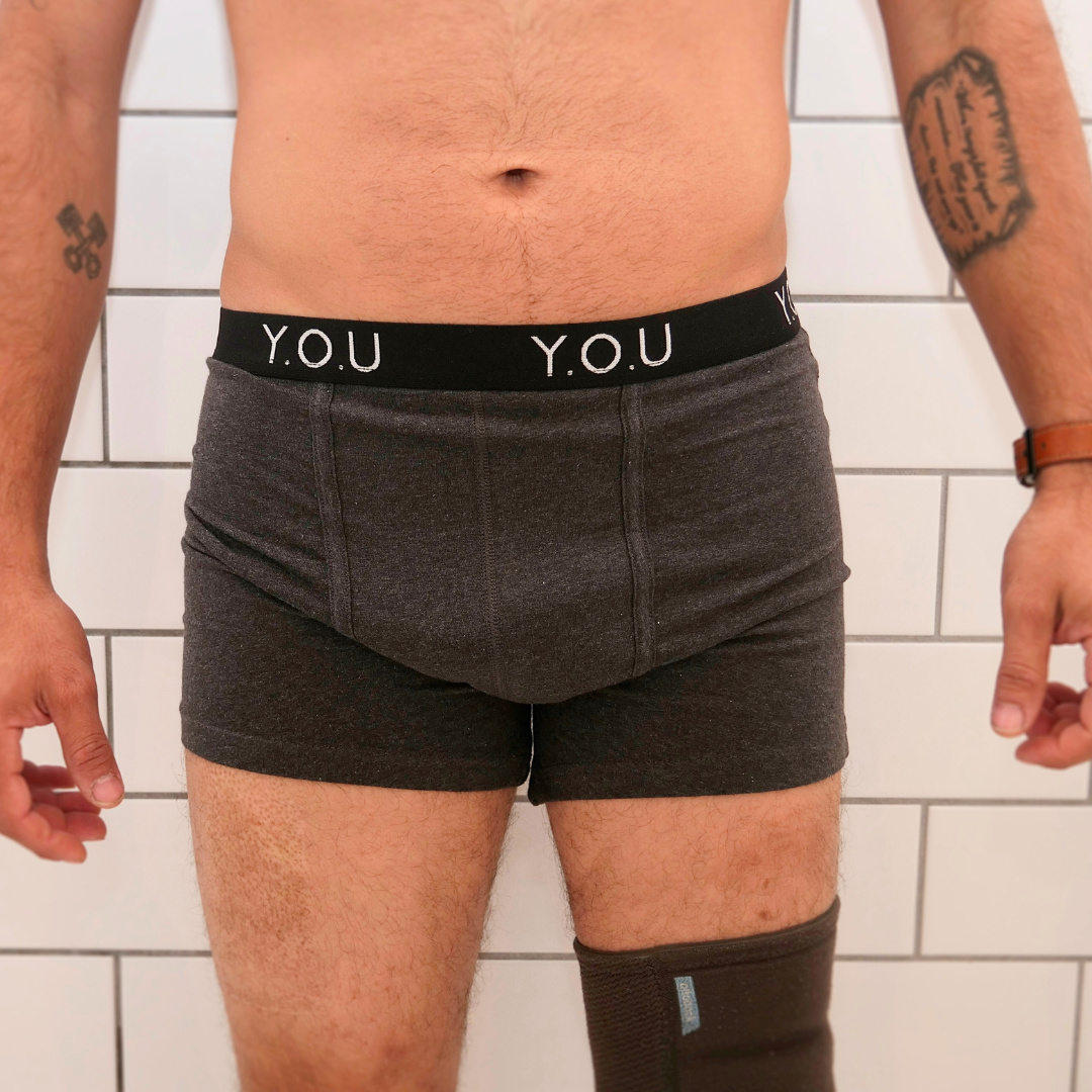 A person wearing dark grey men's mid-length trunks with "Y.O.U" written on the waistband is standing in front of a tiled background. They have arm tattoos and a leg brace on the left thigh, with their arms relaxed at their sides.