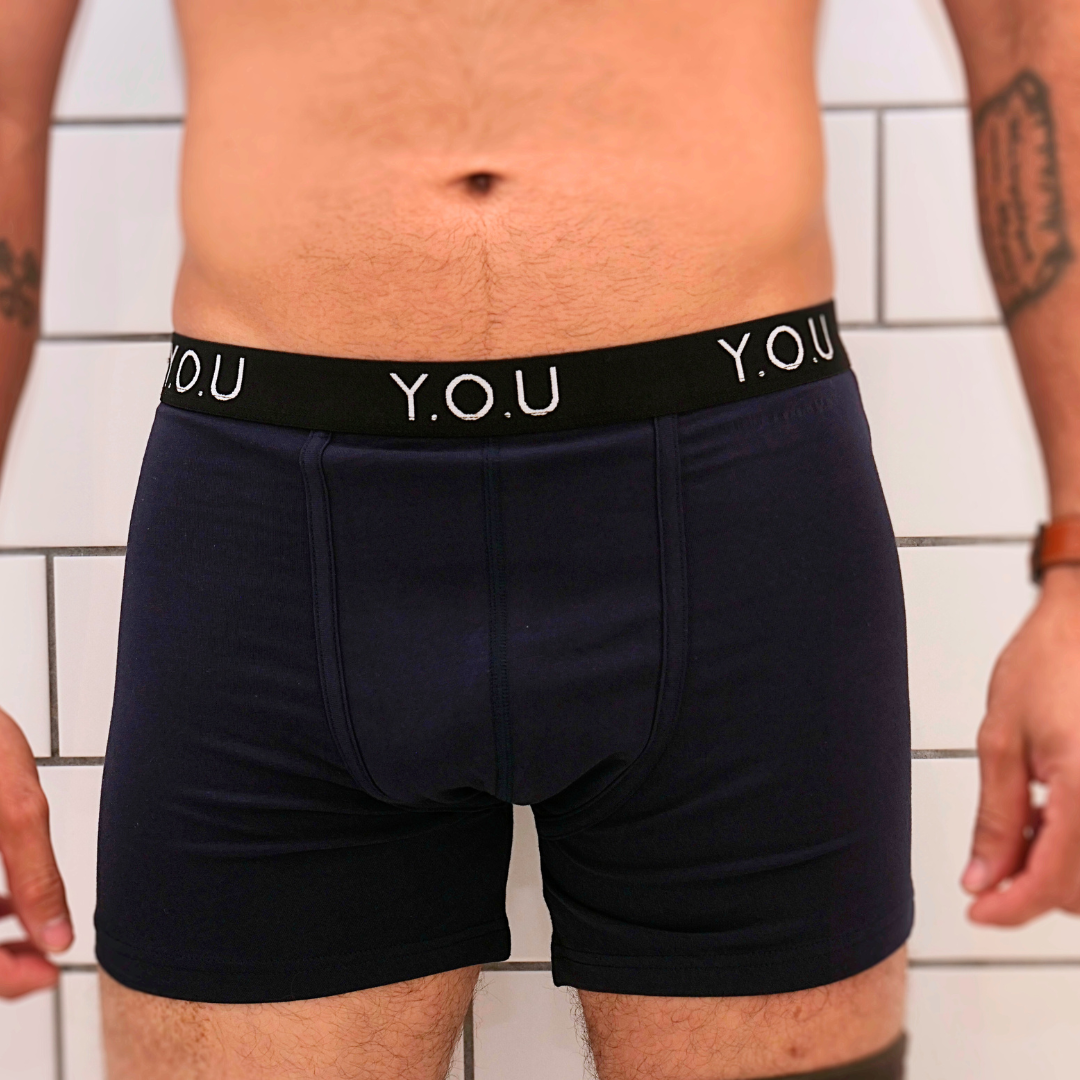 A person stands against a white tiled wall, wearing dark navy blue men’s mid-length trunks with a black waistband that reads "Y.O.U" in white letters. Their wrists are adorned with tattoos and a brown wristwatch. The photo captures the torso and upper thigh area.