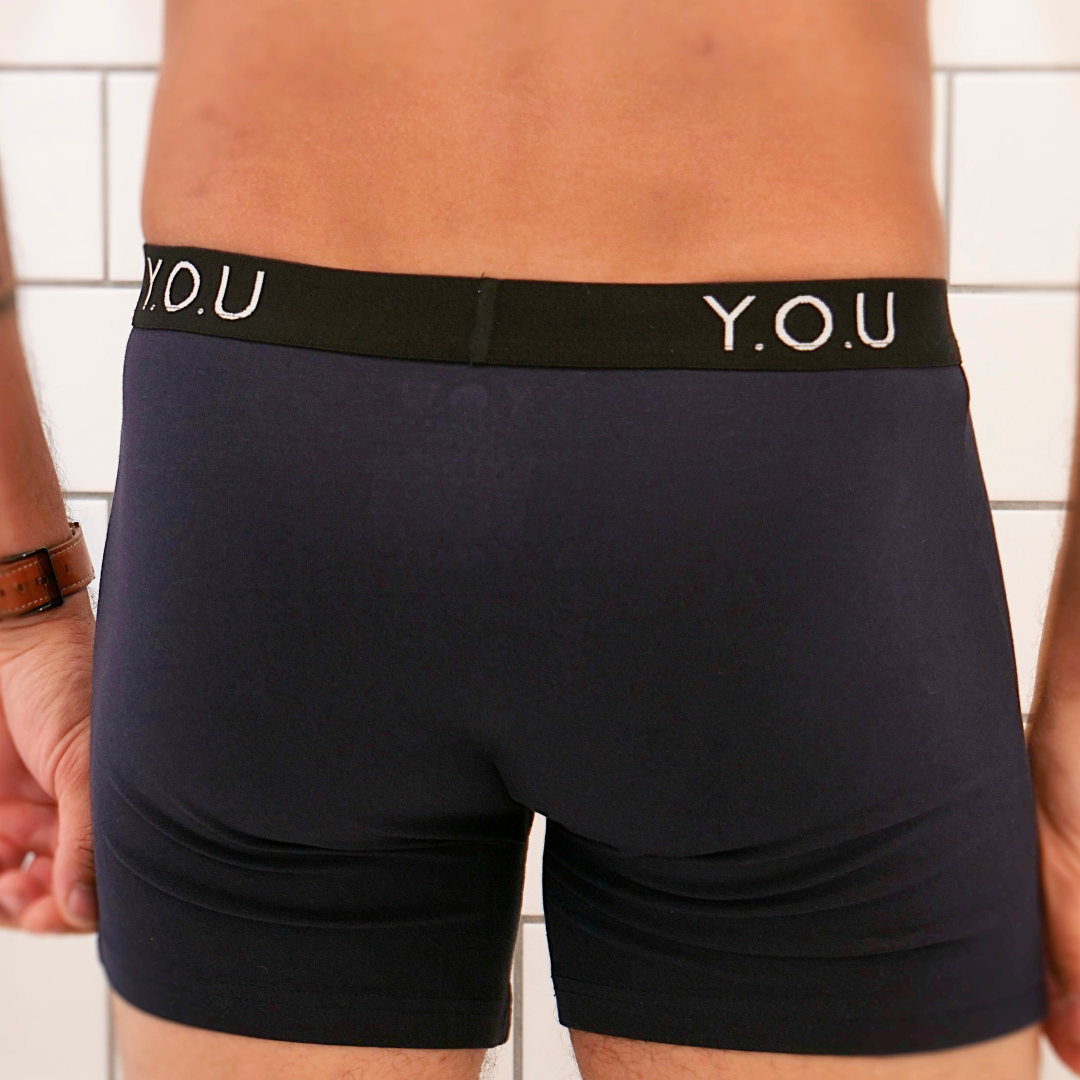 Rear view of a person standing, wearing dark navy blue men's mid length trunks with a black waistband that has the letters "Y.O.U" in white. The background features white subway tiles. 