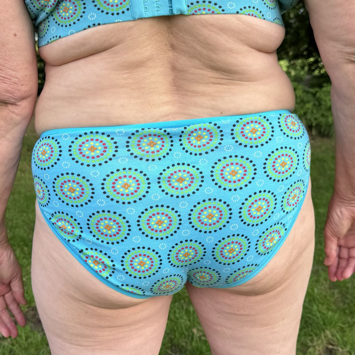 A close up photo of a model wearing the YOU Underwear mid rise bikini knickers in the blue mara print. They are standing facing away from the camera with their hands at their sides. We can see from the model's stomach to their upper-thigh. There is a green field in the background.
