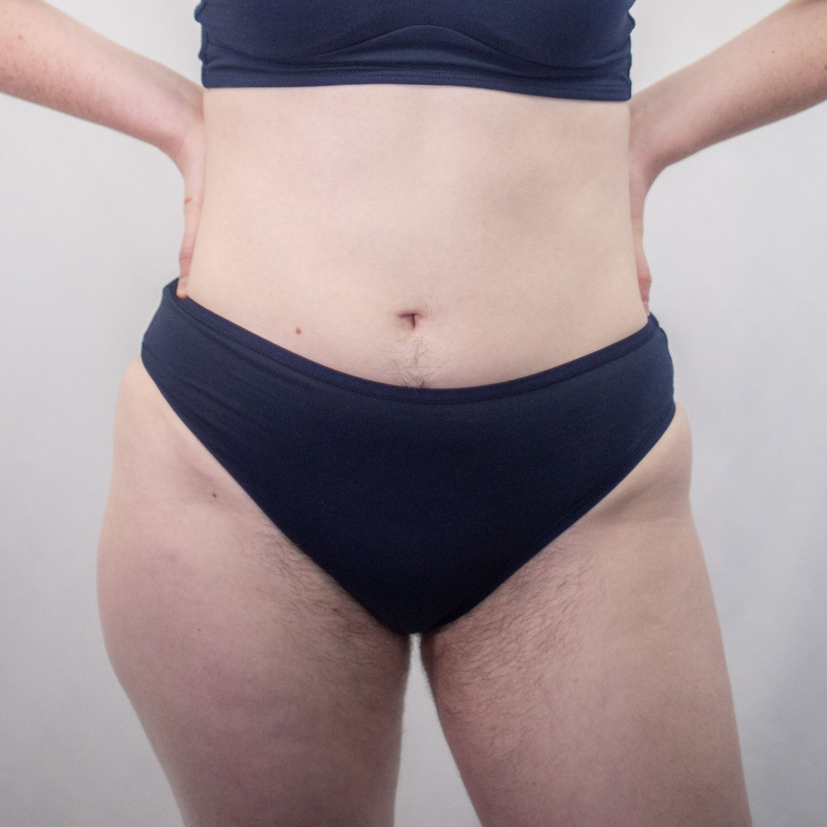 A close up photo of a model wearing the YOU Underwear navy mid rise bikini knicker. The model is standing facing the camera with both their hands on their hips. There is a white background behind them and we can see from the model's stomach to their upper-thigh.