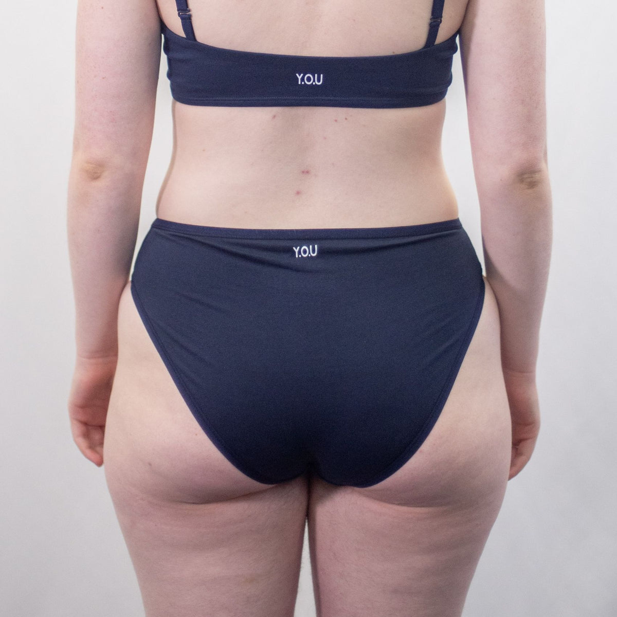 A photo of a model wearing the YOU Underwear navy mid rise bikini knicker and the matching bralette. The model is standing facing away from the camera with their hands at their sides. There is a white background and we can see from the model's shoulder blades to their upper-thigh.