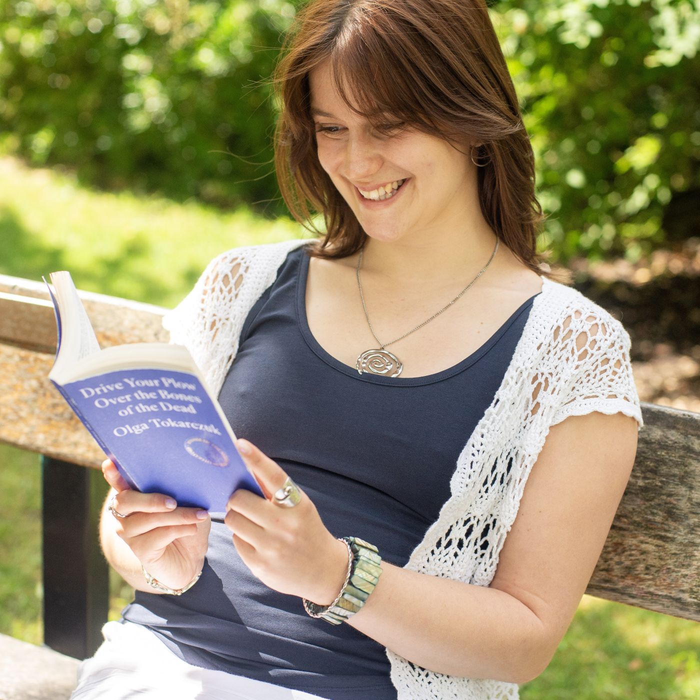 A photo of a model wearing the YOU Underwear vest in navy She is sitting on a bench and smiling at the book she is reading. The background is a green park.