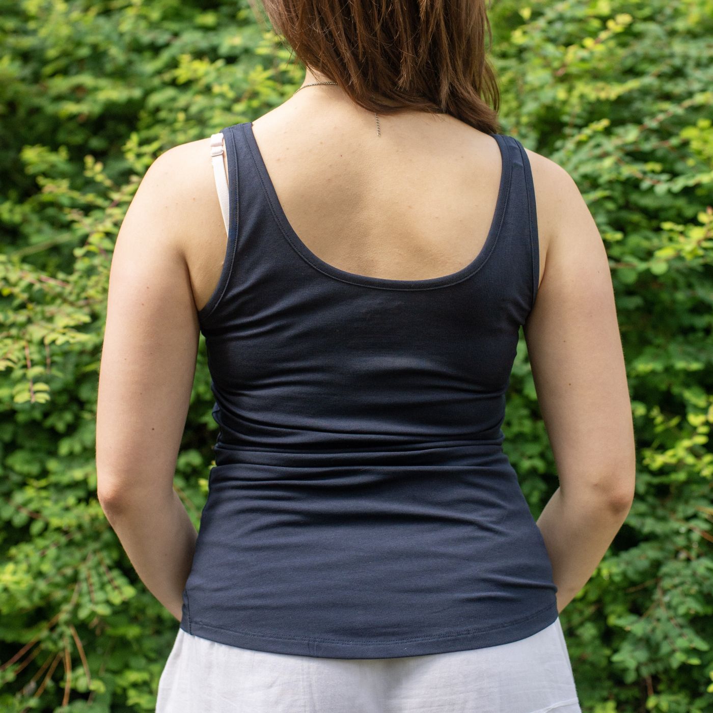 A close up of a model wearing the YOU Underwear navy vest. She is facing away from the camera, with her hands at her sides. She is standing in front of green bush.