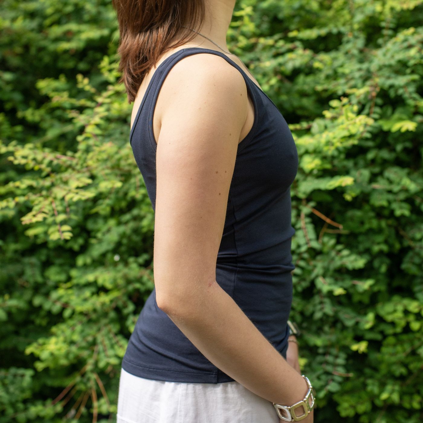 A close up of a model wearing the YOU Underwear navy vest. She is standing side on to the camera, with her hands at her sides. She is standing in front of green bush.