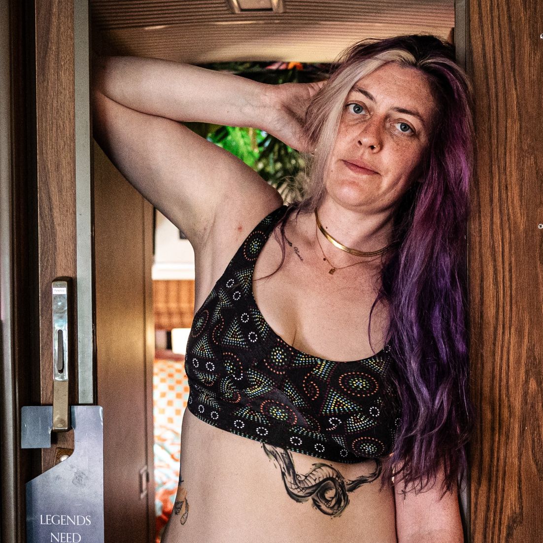 A photo of a model wearing the YOU Underwear non wired bra in black mara. They are standing facing the camera, stood in a door way. They are leaning against the door frame with one arm behind their head. The photo is cropped so we can see to the model's waist.