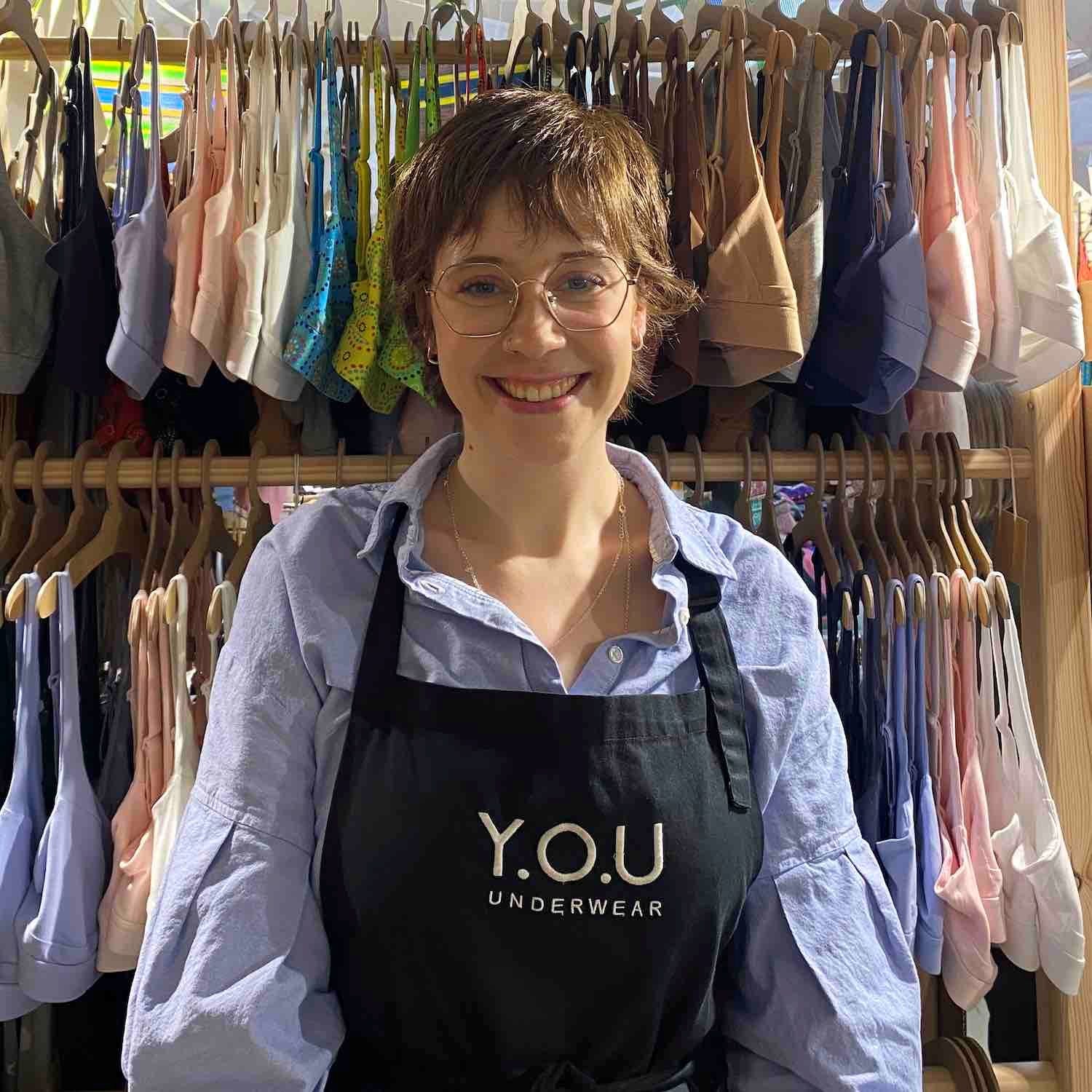 A photo of a YOU Underwear employee. They are facing the camera and smiling. There is a large display of YOU Underwear bras in the background.