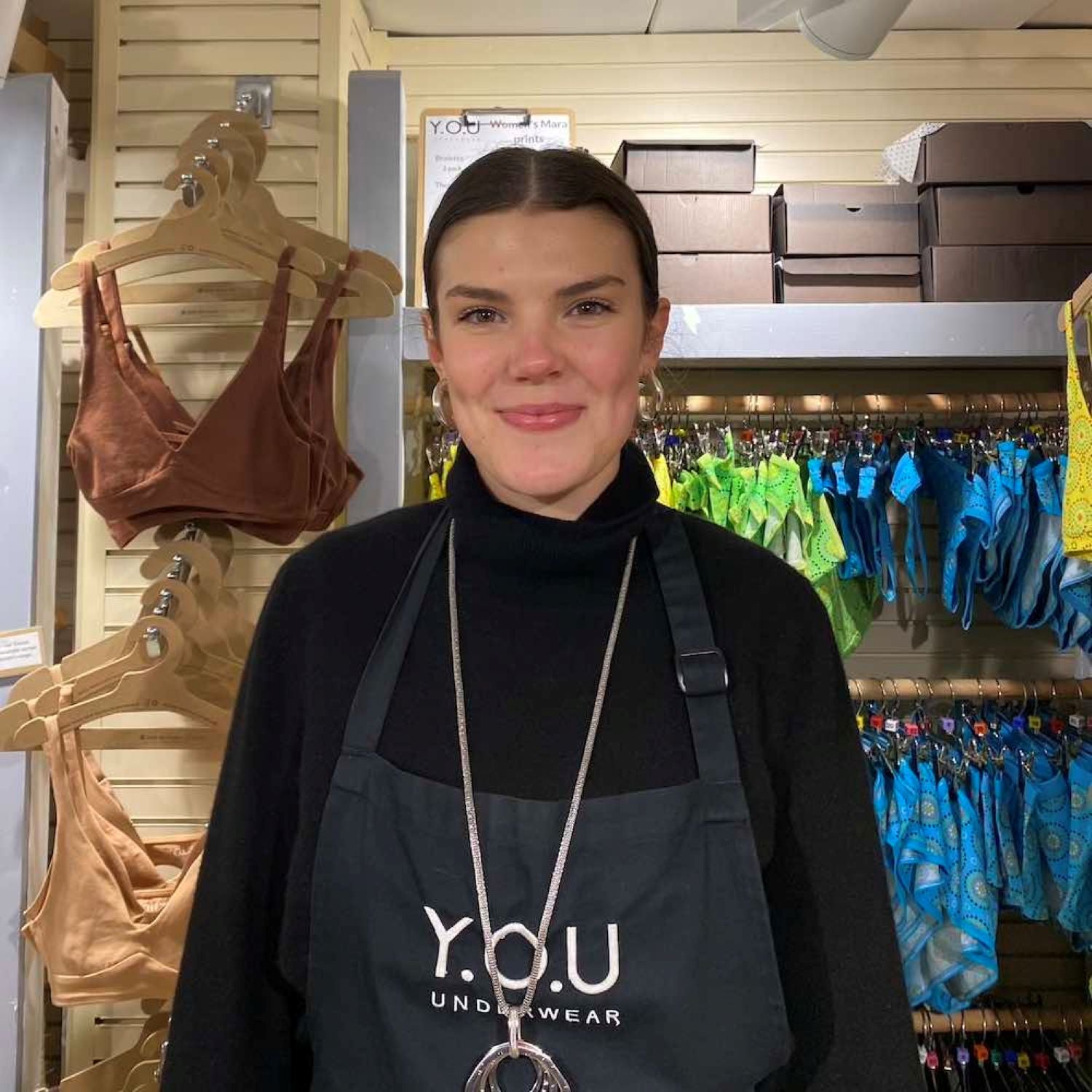 A photo of a YOU Underwear employee. They are facing the camera and smiling. There is a display of YOU Underwear bras in the background.