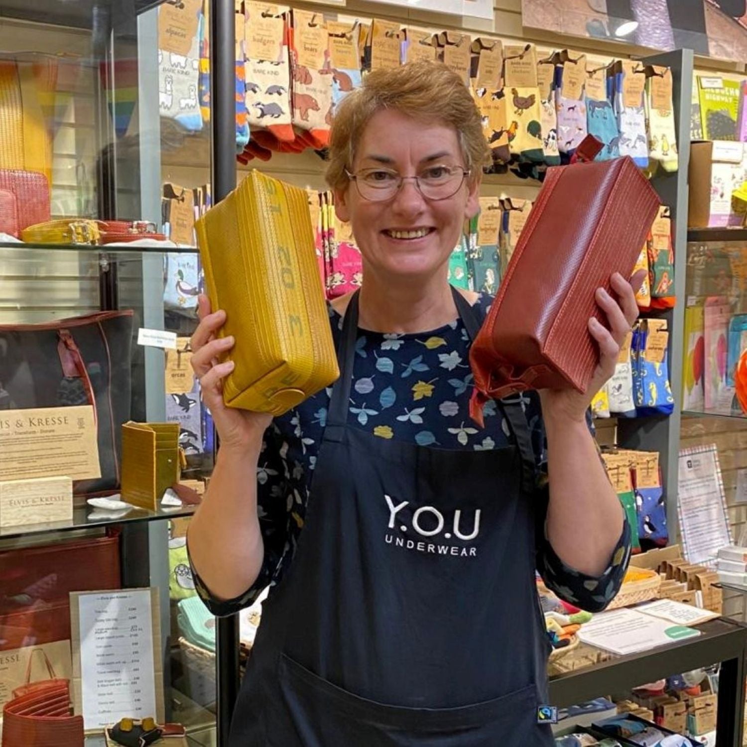 A photo of a YOU Underwear employee. They are facing the camera and smiling whilst holding up two Elvis & Kresse wash bags, one red, one yellow. There is a a large sock display in the background.