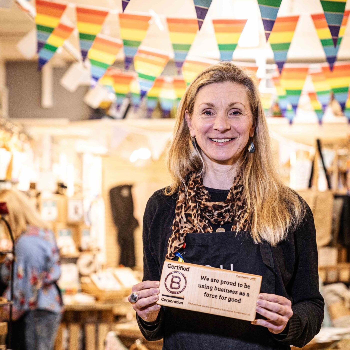 A photo of the YOU Underwear founder, Sarah Jordan. They are facing the camera and smiling whilst holding a wooden plaque which reads, 'Certified B Corp, We are proud to be using business ad a force for good.' There is a large display of YOU Underwear bras in the background.