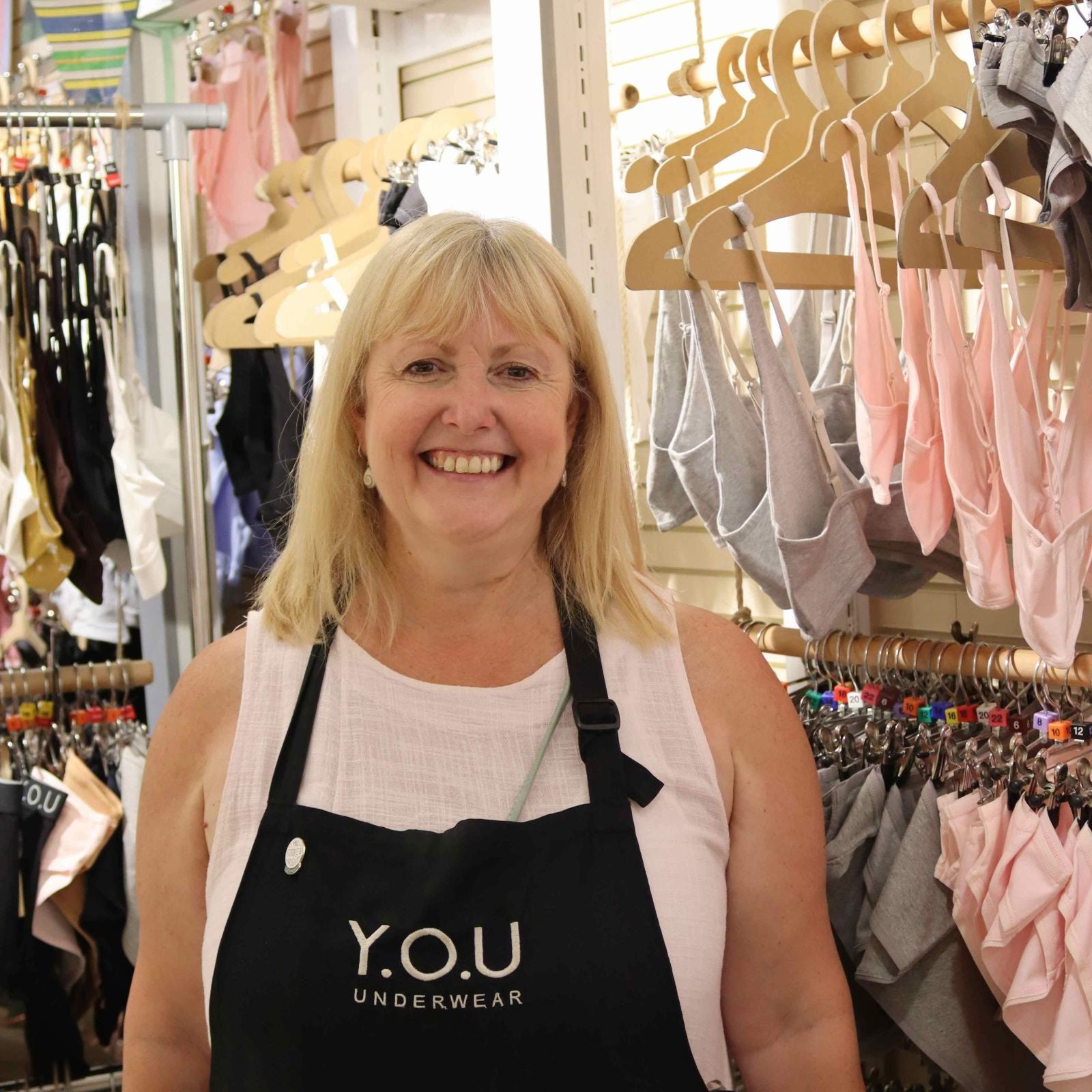 A photo of a YOU Underwear employee. They are facing the camera and smiling. There is a large display of grey and light pink YOU Underwear bras and pants in the background.