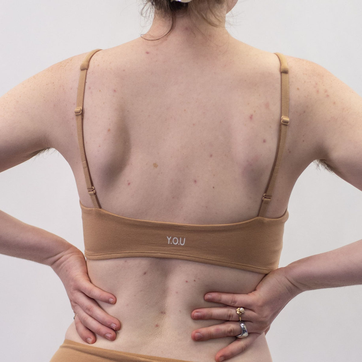 A photo of a model wearing the YOU Underwear padded bralette in almond. The model is standing facing away from the camera with both hands on their hips. The photo is cropped so we can see from the model's shoulders to their waist. There is a white background.