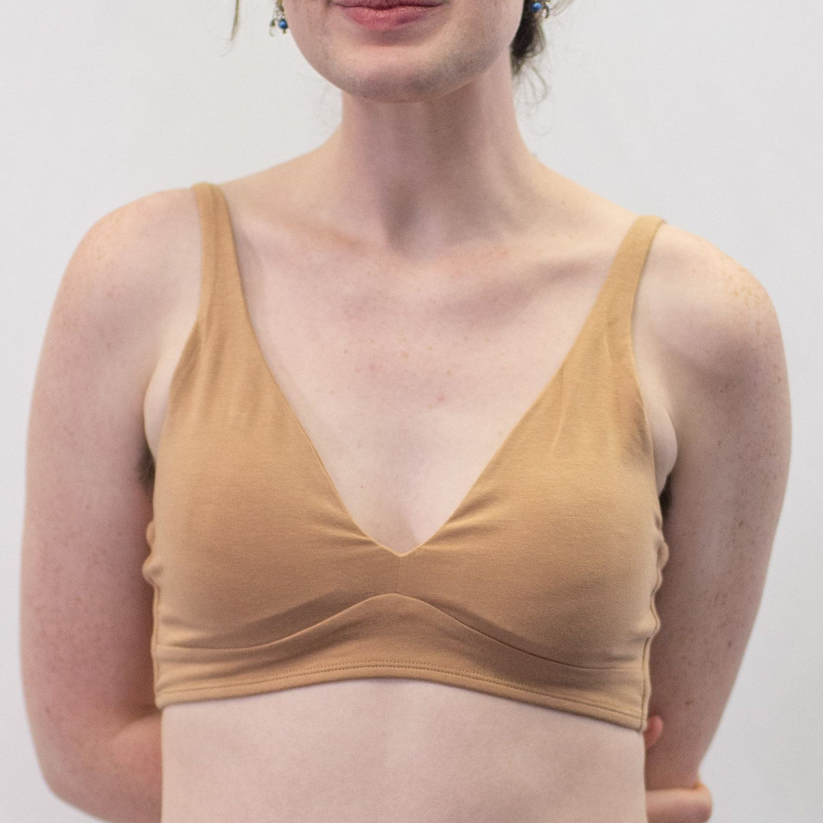 A photo of a model wearing the YOU Underwear padded bralette in almond. The model is standing facing the camera with one hand behind her back and the other at her side. The photo is cropped so we can see from the model's shoulders to their waist. There is a white background.