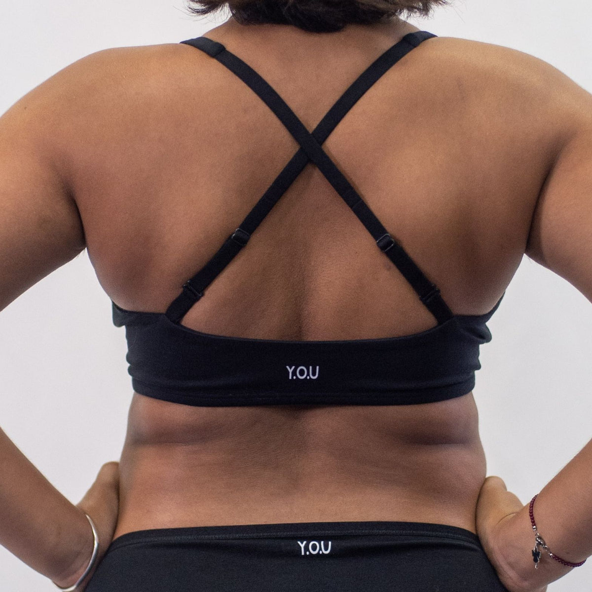 A photo of a model wearing the YOU Underwear padded bralette in black. They are standing facing away from the camera with their hands on their hips. The photo is cropped so we can only see to the model's waist. The photo shows off the bralette which has its straps crossed over to show another style of wear. There is a white background.