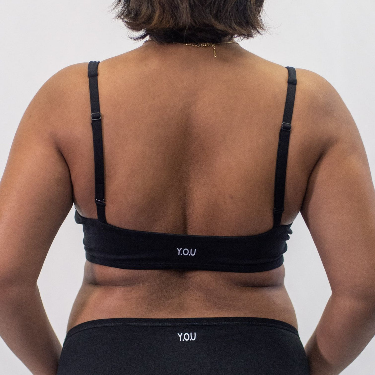 A close up photo of a model wearing the YOU Underwear padded bralette in black. They are standing facing away from the camera with their hands at their sides. The photo is zoomed in so we can see from the model's shoulders to their hips. They are also wearing black YOU Underwear bikini knickers. There is a white background.