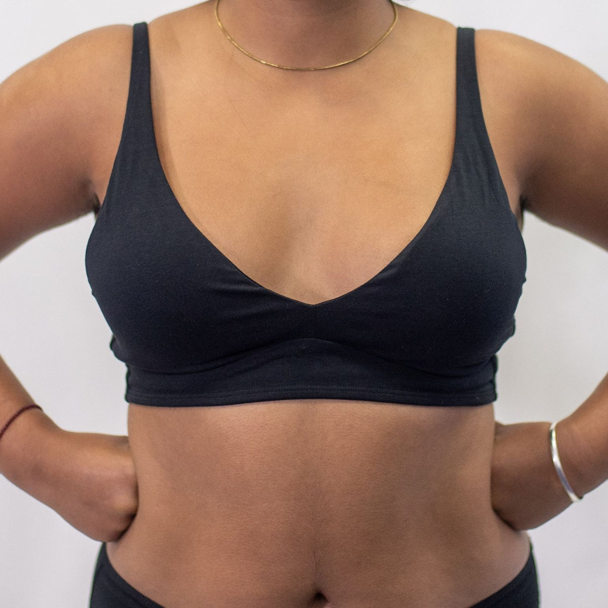 A close up photo of a model wearing the YOU Underwear padded bralette in black. They are standing facing the camera with their hands on their hips. The photo is zoomed in so we can see from the model's shoulders to their waist. There is a white background.