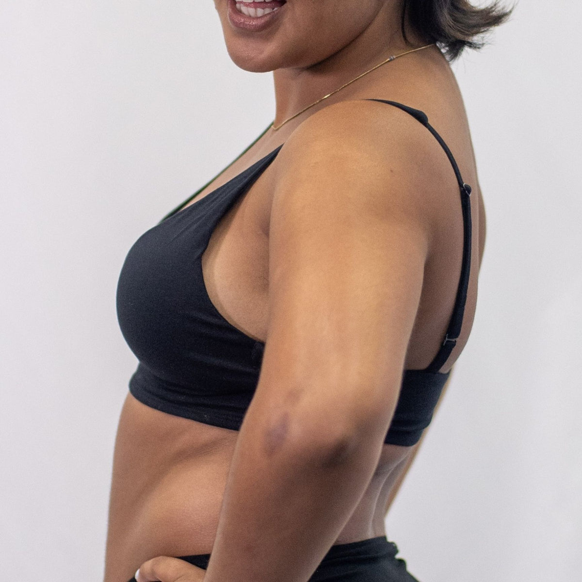 A photo of a model wearing the YOU Underwear padded bralette in black. They are standing side on to the camera with their hands on their hips. The photo is cropped so we can only see to the model's waist. There is a white background.
