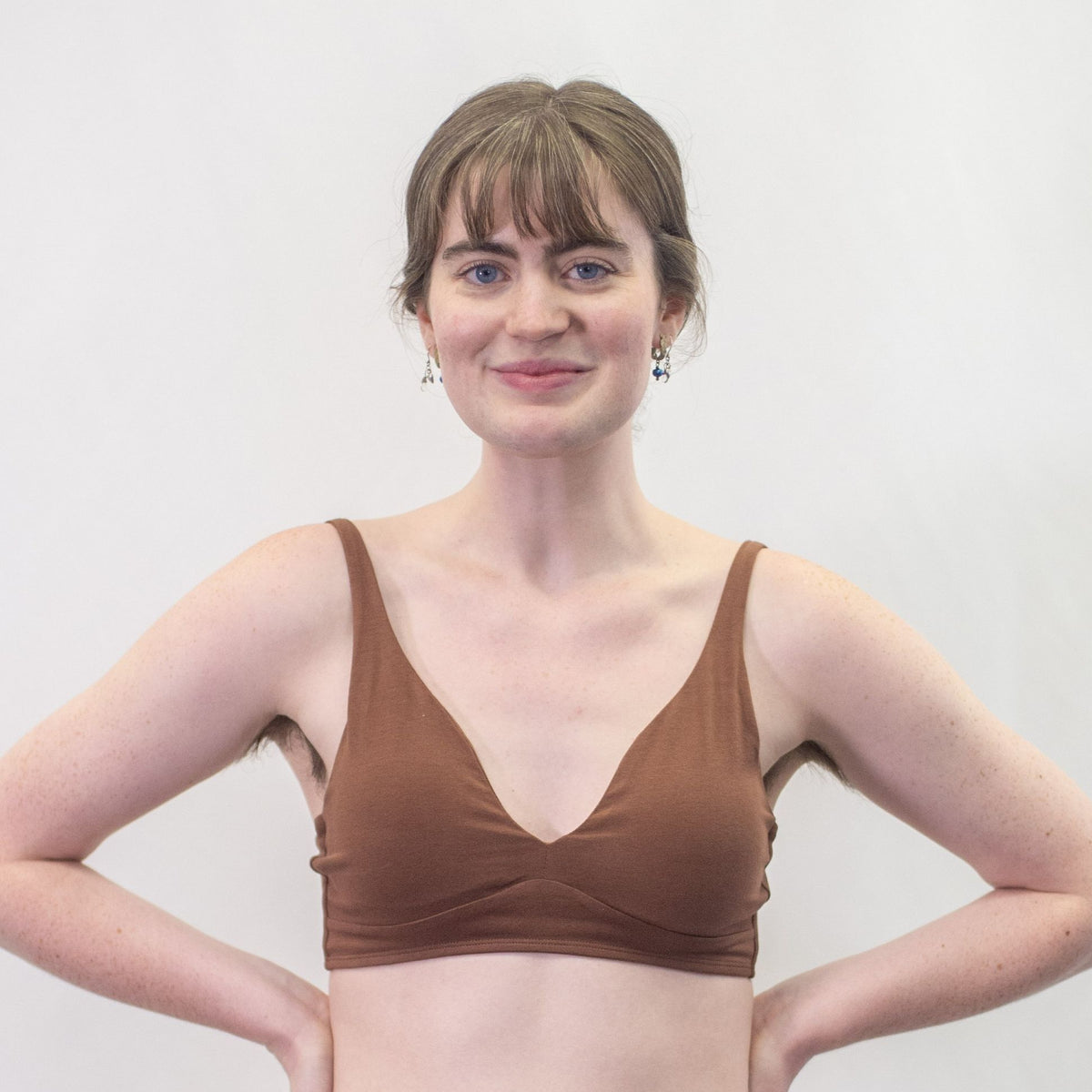 A photo of a model wearing the YOU Underwear padded bralette in chestnut. She is standing facing the camera with both hands on her hips. The photo is cropped so we can see to the model's mid-stomach. There is a white background.