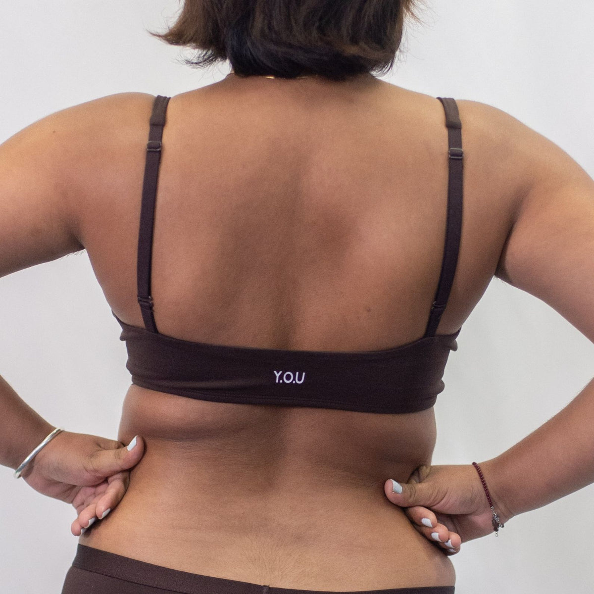A photo of a model wearing the YOU Underwear padded bralette in cocoa. She is standing facing away from the camera with both hands on her hips, smiling. The photo is cropped so we can see from the model's shoulder to their hips. There is a white background.