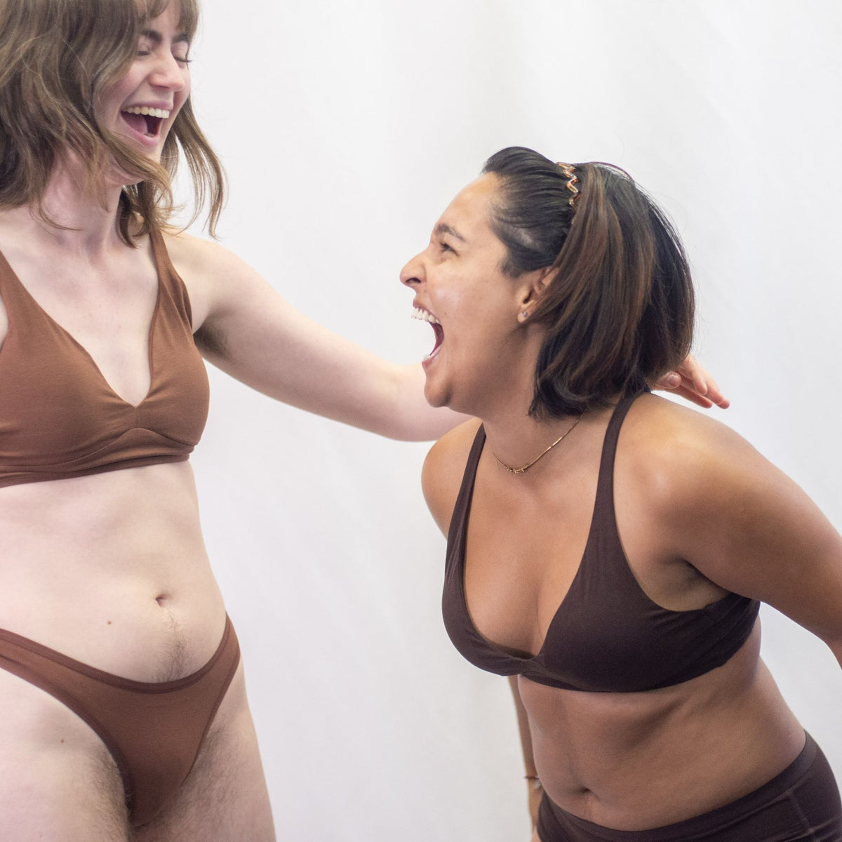 A photo of two models, one wearing the YOU Underwear padded bralette and thong in chestnut and the other wearing a cocoa padded bralette and matching boyshorts. The photo is candid and they are laughing together. There is a white background.