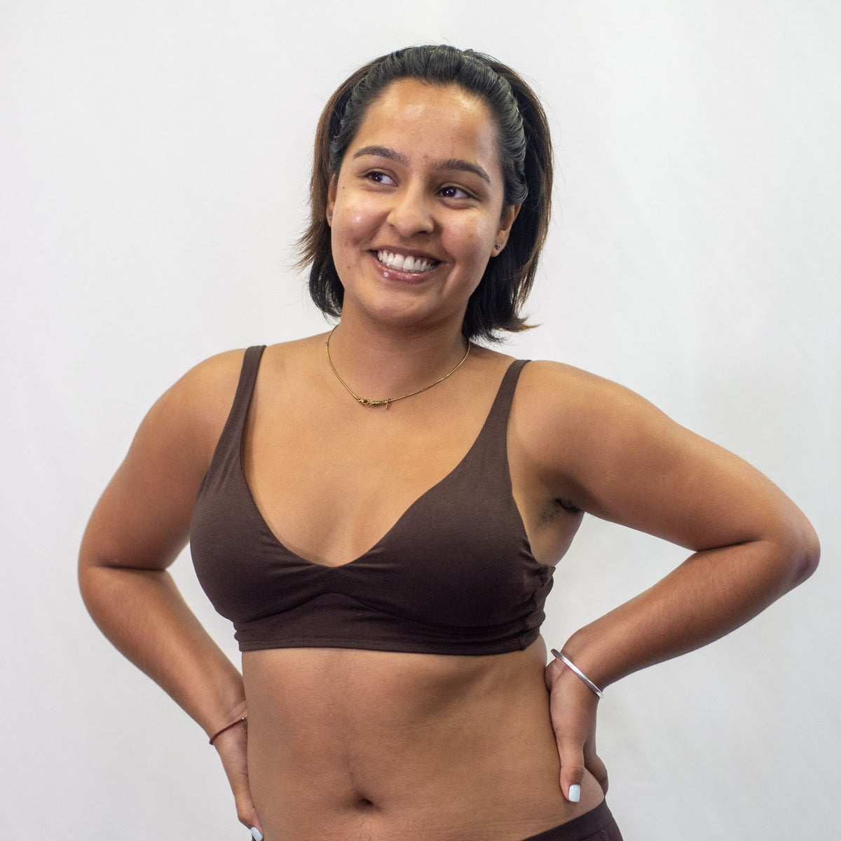 A photo of a model wearing the YOU Underwear padded bralette in cocoa. She is standing facing the camera with both hands on her hips, smiling. The photo is cropped so we can see to the model's hips. There is a white background.