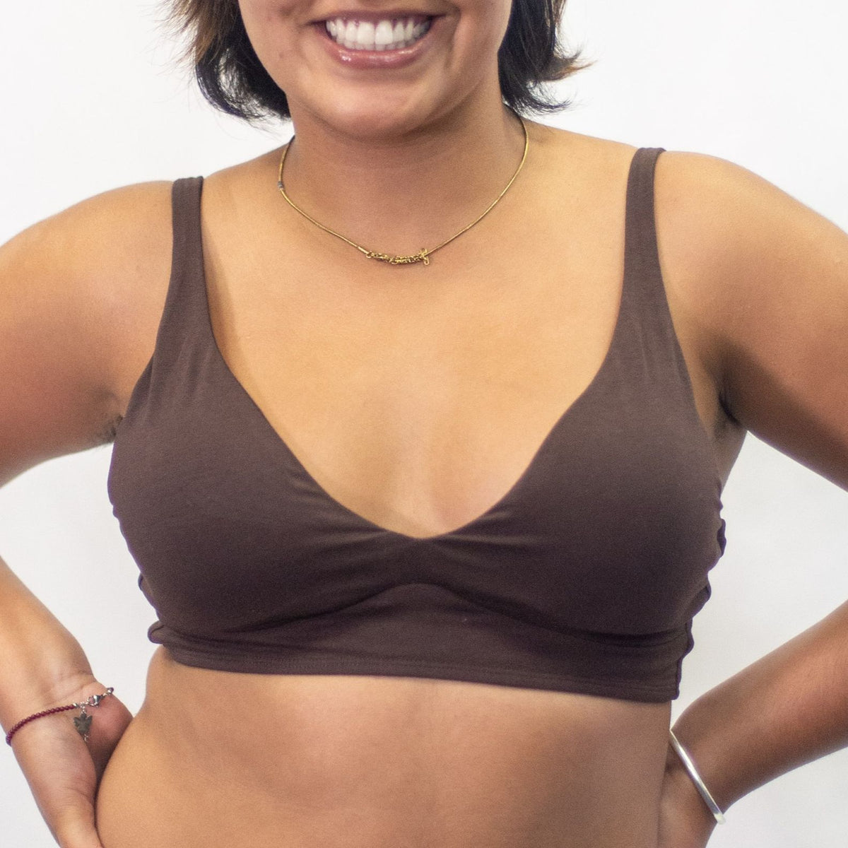 A photo of a model wearing the YOU Underwear padded bralette in cocoa. She is standing facing the camera with both hands on her hips, smiling. The photo is cropped so we can see from the model's jaw to their mid-stomach. There is a white background.