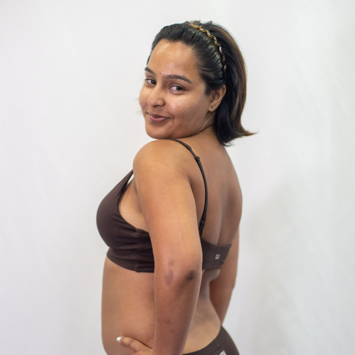 A photo of a model wearing the YOU Underwear padded bralette in cocoa. She is standing side on to the camera with both hands on her hips, turning her head and smiling at the lens. The photo is cropped so we can see to the model's hips. There is a white background.