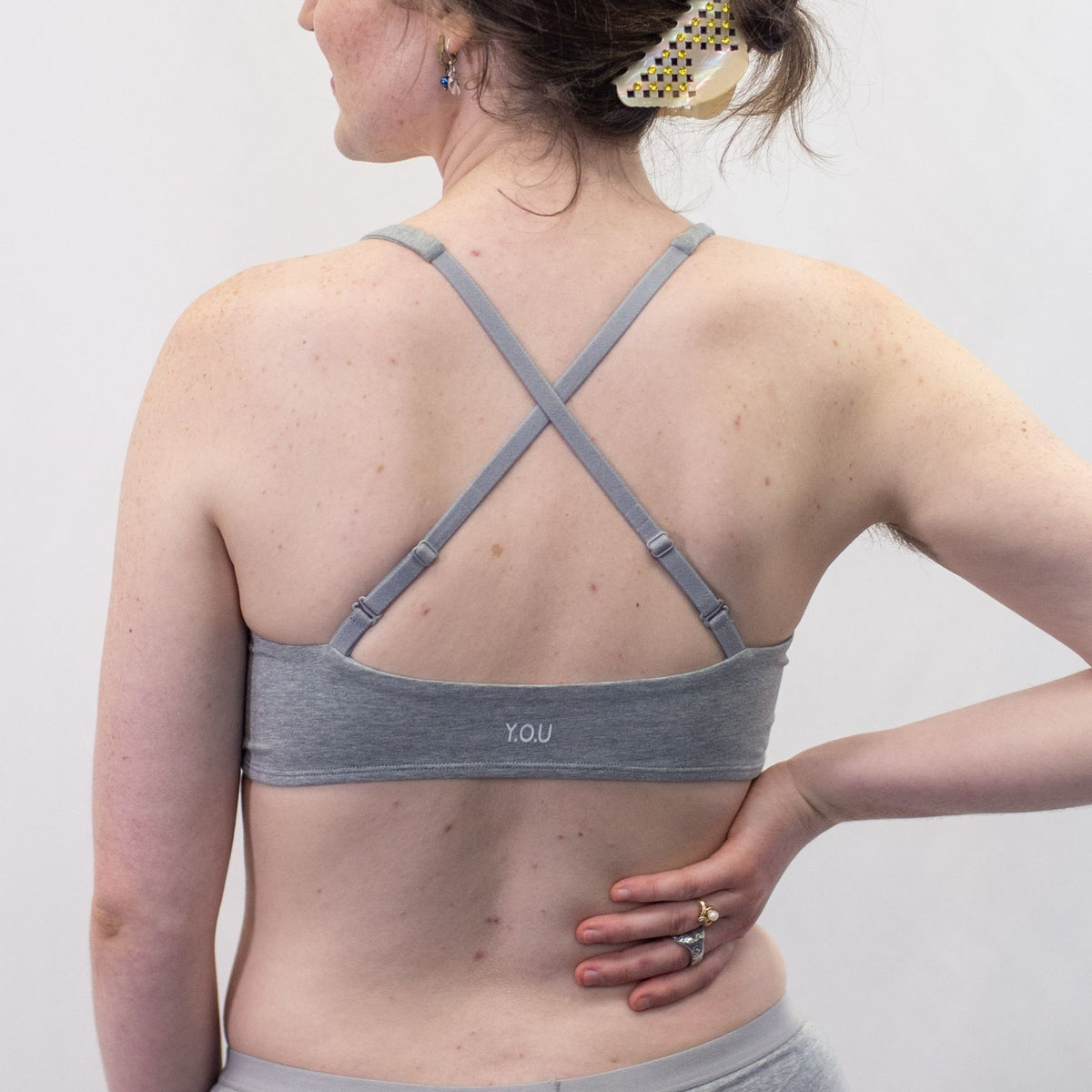 A photo of a model wearing the YOU Underwear padded bralette in grey. They are standing facing away from the camera with their hands on their hips. The photo is zoomed in so we can see from the model's shoulders to their mid-stomach. The bralette straps are crossed to show another style of wear. There is a white background.