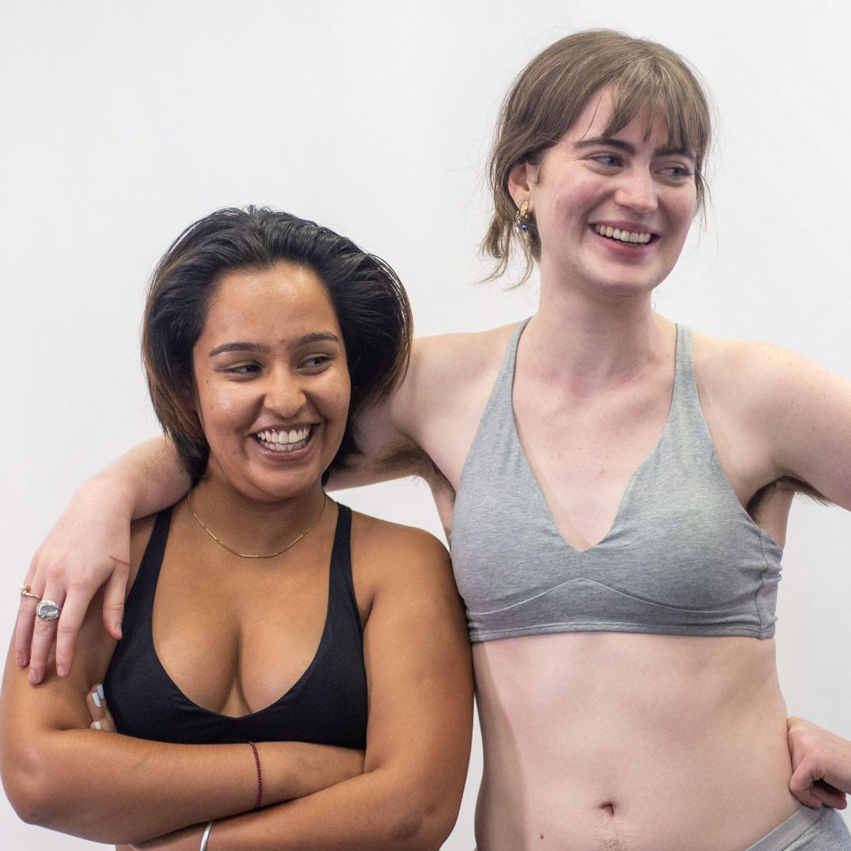 A photo of two models, one wearing the YOU Underwear padded bralette in grey and one in black. They are standing facing the camera, the model in grey with an arm over the other model. They are looking off to the left and laughing. The photo is cropped so we can see to their stomach. There is a white background.
