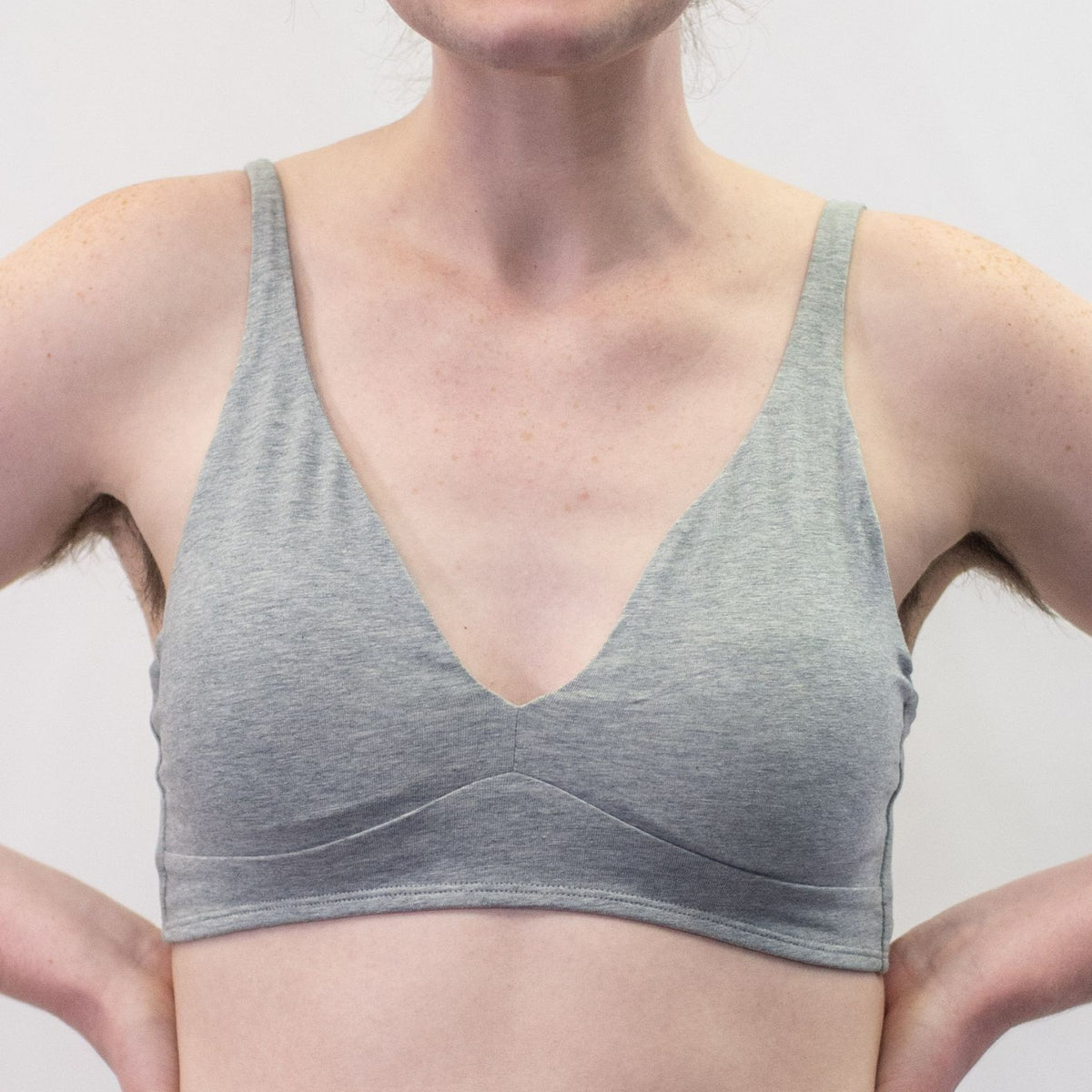 A photo of a model wearing the YOU Underwear padded bralette in grey. They are standing facing the camera with their hands on their hips. The photo is zoomed in so we can see from the model's shoulders to their mid-stomach. There is a white background.