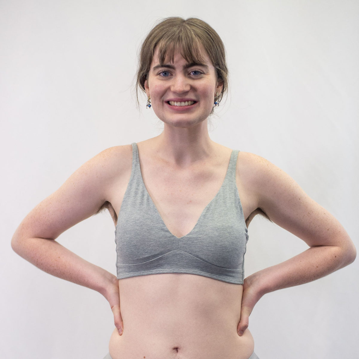 A photo of a smiling woman with brown hair and freckles wearing the YOU Underwear padded bralette in grey. They are standing facing the camera with their hands on their hips. The photo is zoomed in so we can see the model down to just below their belly button. There is a white background.