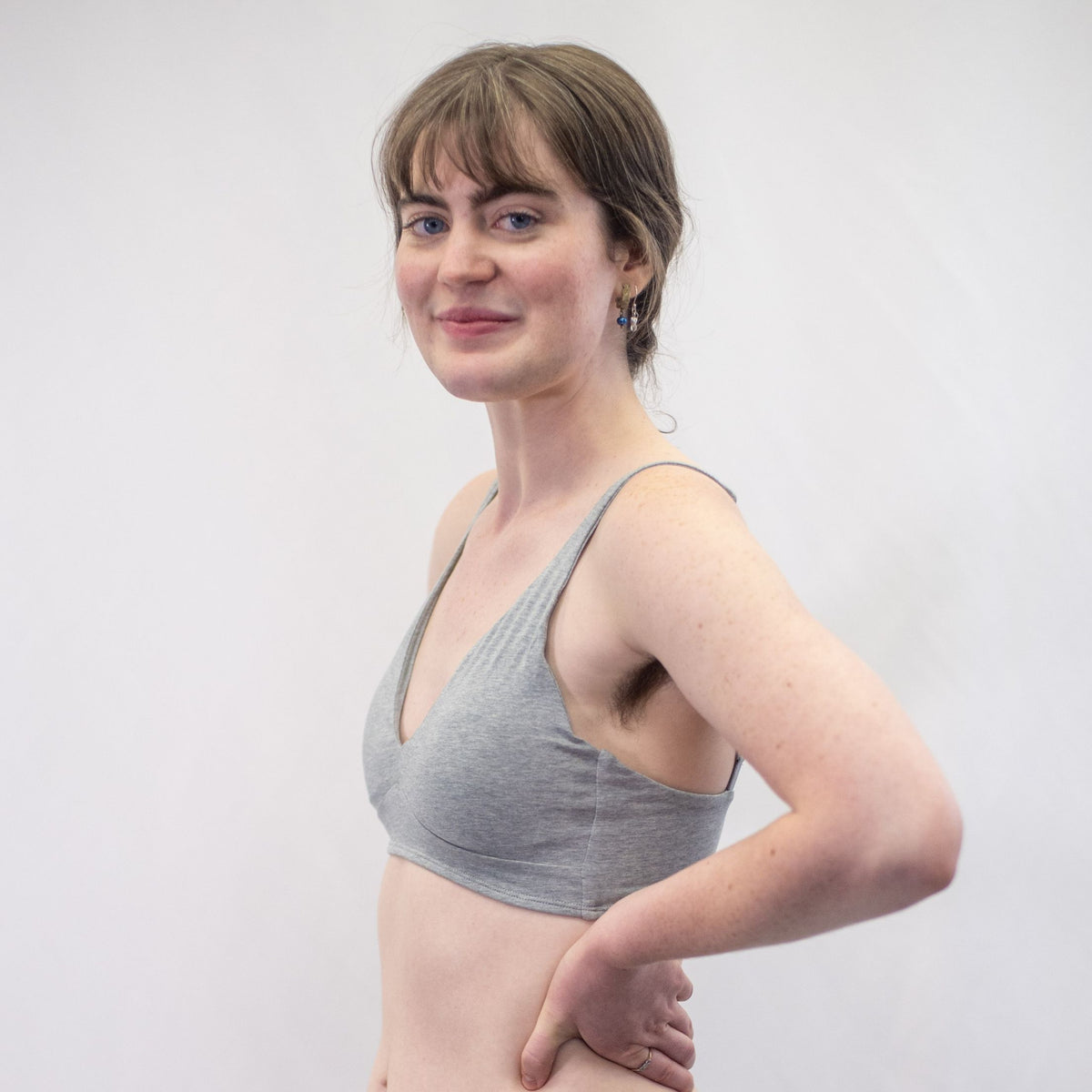A photo of a model wearing the YOU Underwear padded bralette in grey. They are standing side on to the camera with their hands on their hips. The photo is zoomed in so we can see  to their mid-stomach. There is a white background.