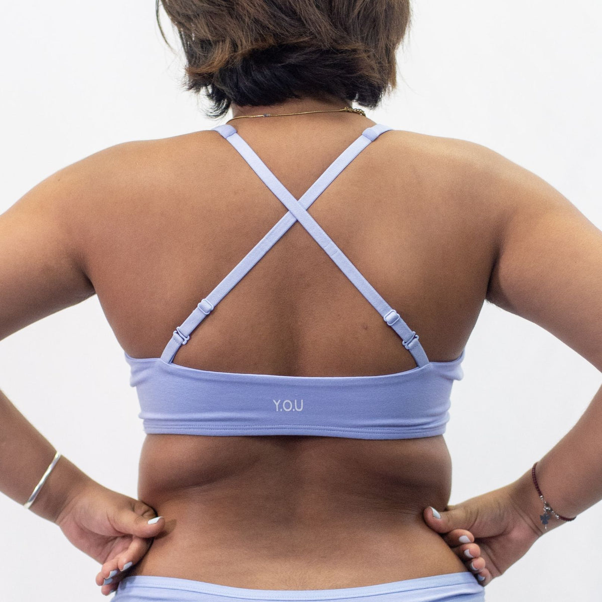A photo of a model wearing the YOU Underwear padded bralette in light blue. She is standing facing away from the camera with both hands on her hips. The photo is cropped so we can see to the model's waist. The straps of the bralette are crossed to show a different style of wear. There is a white background.