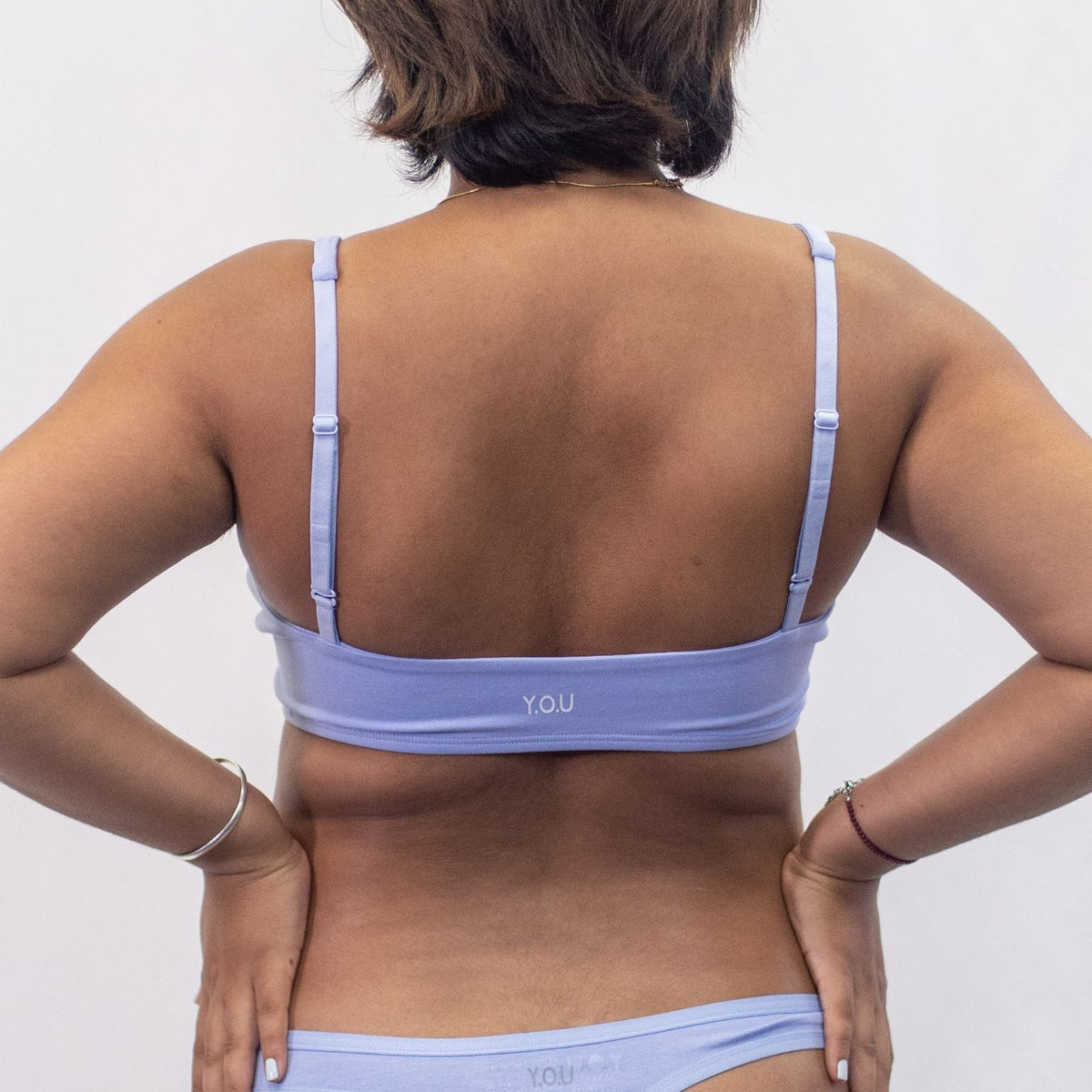 A photo of a model wearing the YOU Underwear padded bralette in light blue. She is standing facing away from the camera with both hands on her hips. The photo is cropped so we can see from the model's neck to their hips. There is a white background.