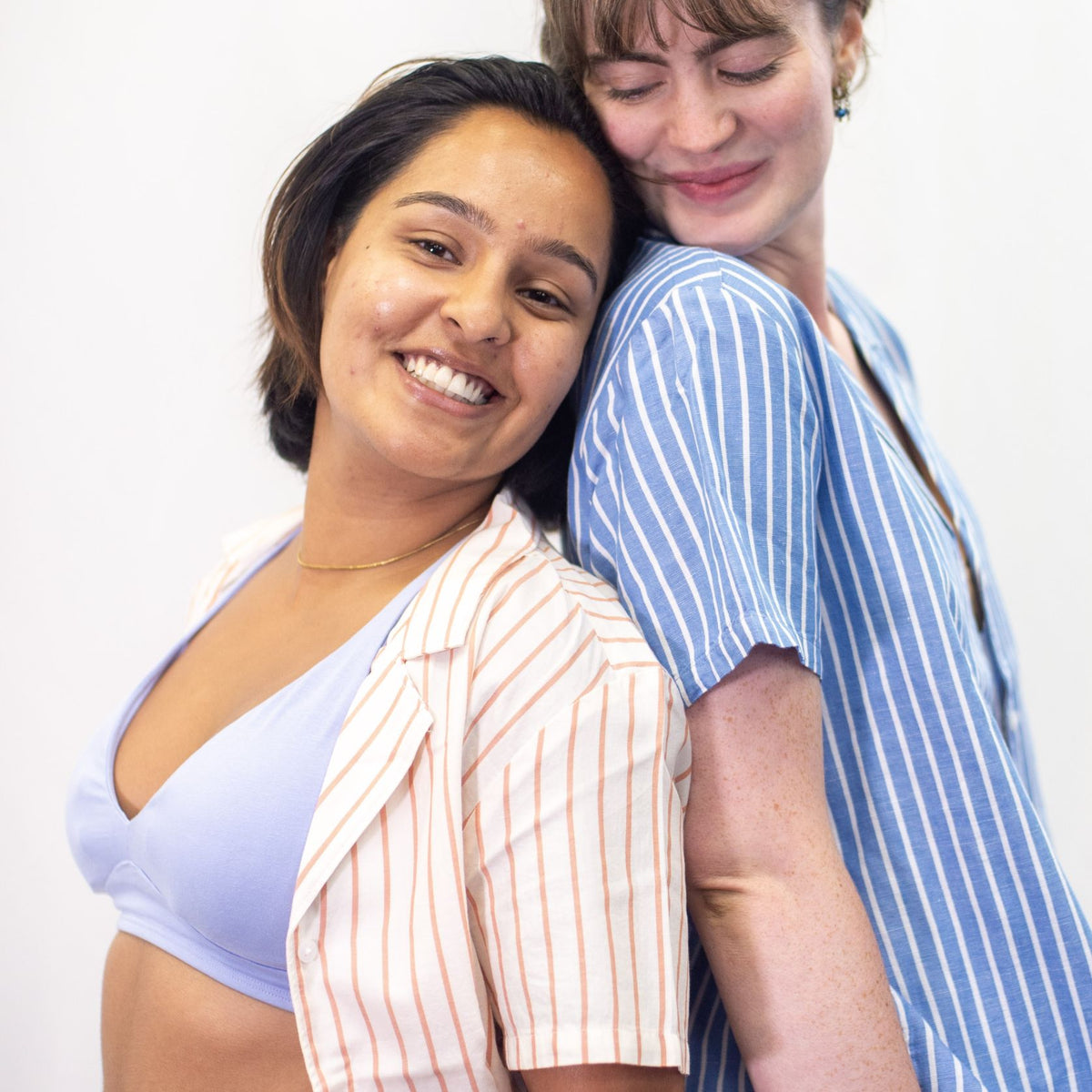 A photo of two models, one is wearing the YOU Underwear padded bralette in light blue paired with a cream and coral striped shirt. The other is wearing a blue and white striped shirt. They are leaning together, back to back and smiling. There is a white background.