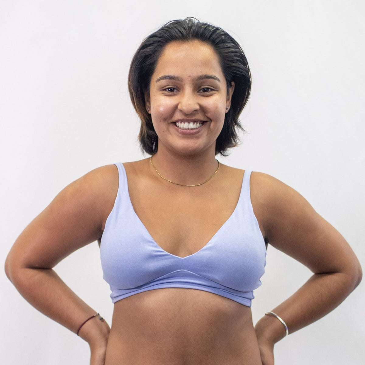 A photo of a model wearing the YOU Underwear padded bralette in light blue. She is standing facing the camera with both hands on her hips and smiling at the camera. The photo is cropped so we can see to the model's waist. There is a white background.