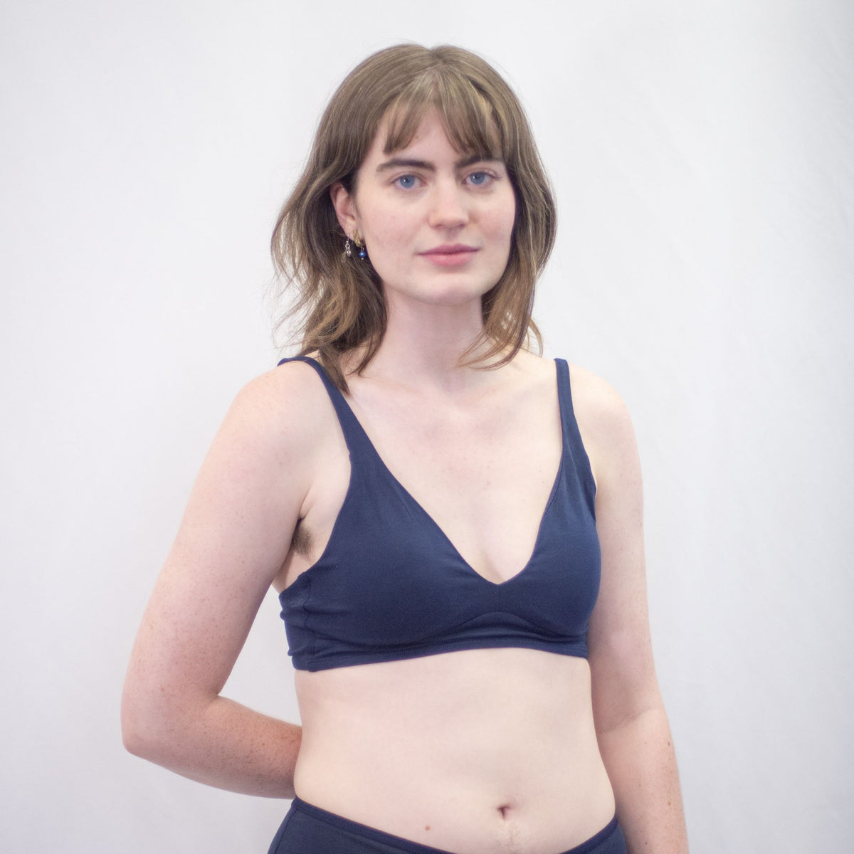 A photo of a model wearing the YOU Underwear padded bralette in navy blue. She is standing facing the camera with one hand behind her back and one at her side. There is a white background and the photo is cropped so we can see to the model's waist.