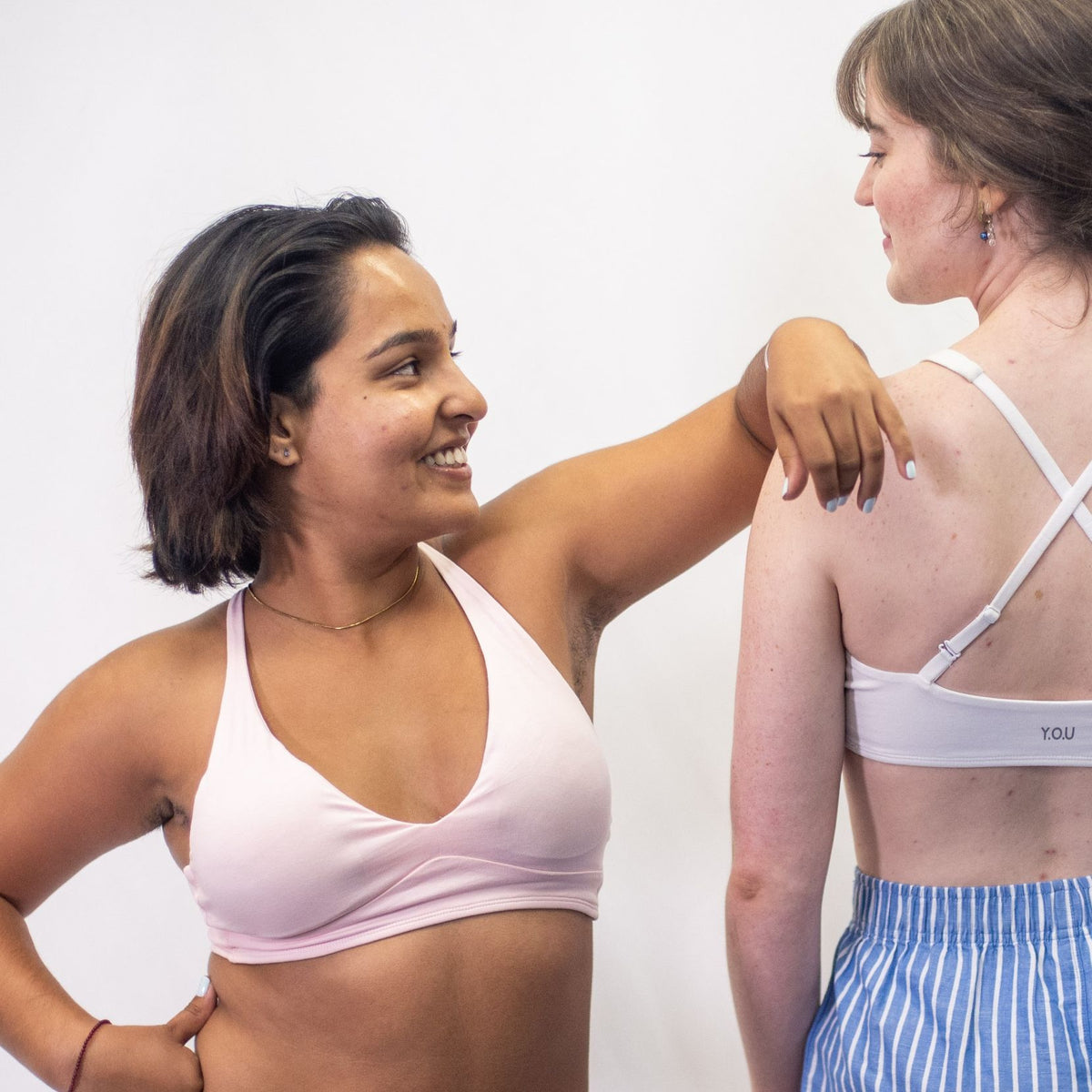 A photo of a model wearing the YOU Underwear padded bralette in pink. There is another model standing next to her, wearing the white bralette with the straps crossed. The model in pink is standing facing the camera, with her left arm on the other model's shoulder. They are looking at each other and smiling. There is a white background.