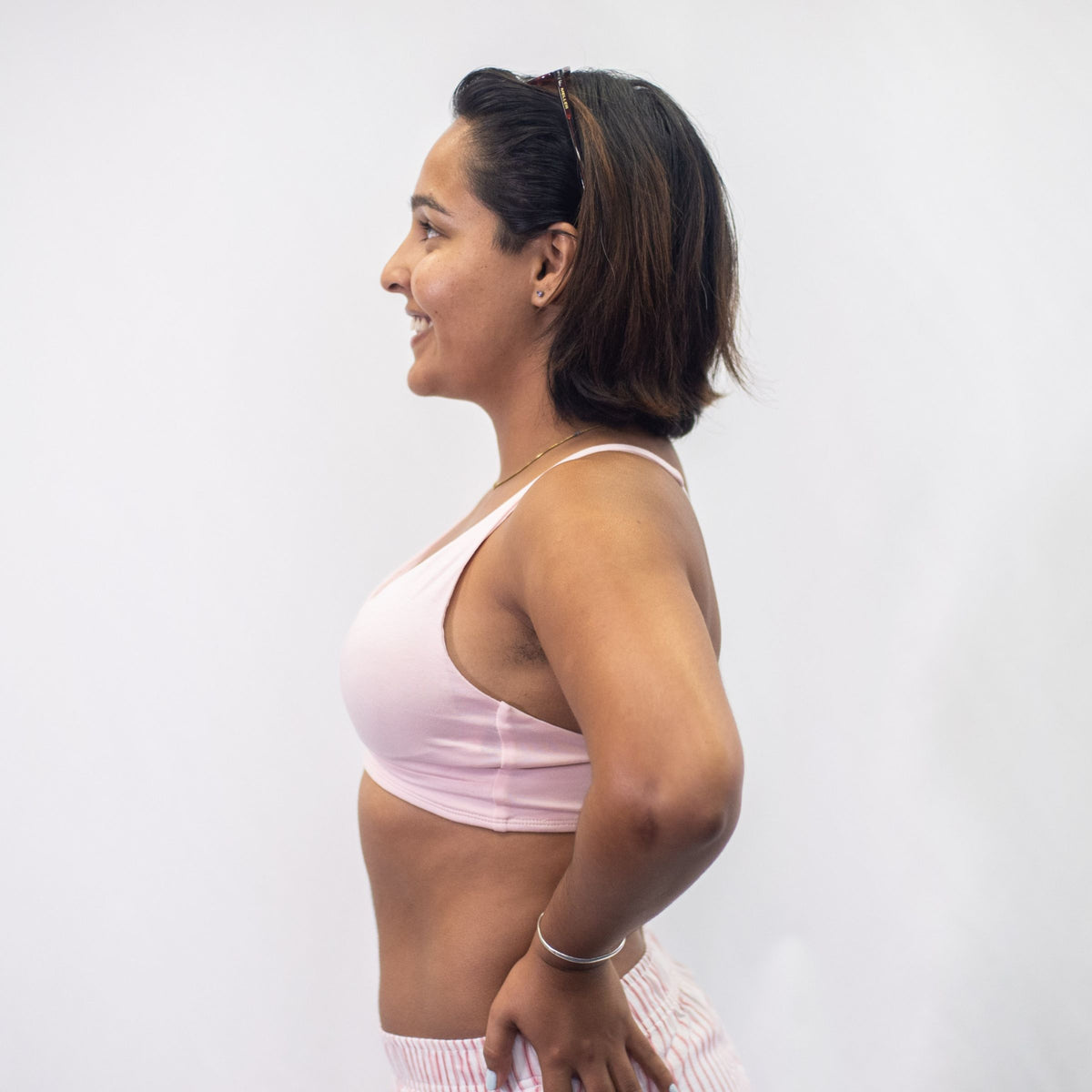 A photo of a model wearing the YOU Underwear padded bralette in pink. She is standing facing side on to the camera with both hands on her hips. The photo is cropped so we can see to the model's hips. There is a white background.
