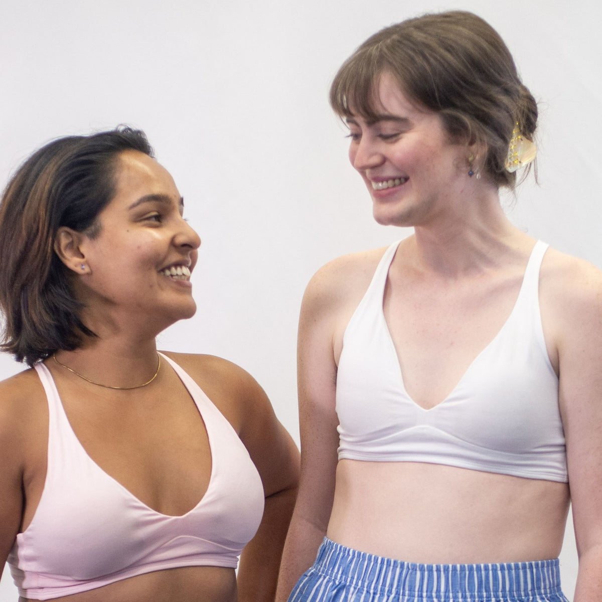 A photo of two models, one wearing the YOU Underwear padded bralette in white and one wearing it in pink. They are both standing facing the camera and they are smiling at each other. There is a white background.