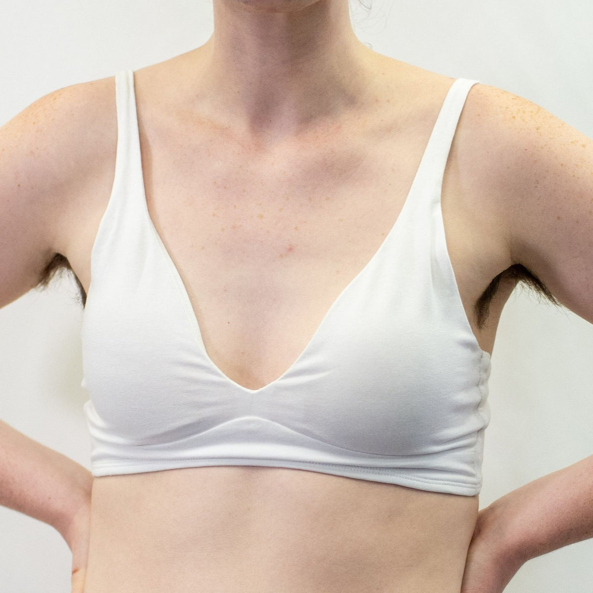 A close up photo of a model wearing the YOU Underwear bralette in white. They are standing facing the camera with both hands on their hips. The photo is zoomed in so we can see from the model's shoulders to their mid-stomach. The background is white.