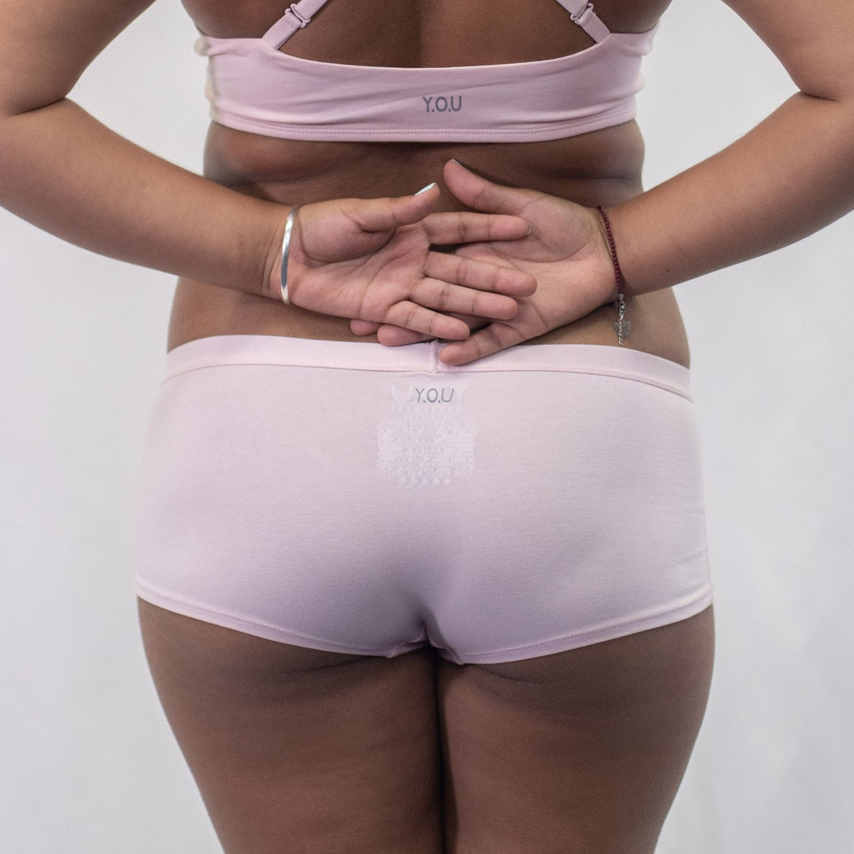 A photo of a model wearing the YOU Underwear boyshorts in pink. They are standing facing away from the camera with their hands held behind their back. The photo is zoomed in so we can see from the model's stomach to their upper-thigh. The background is white.