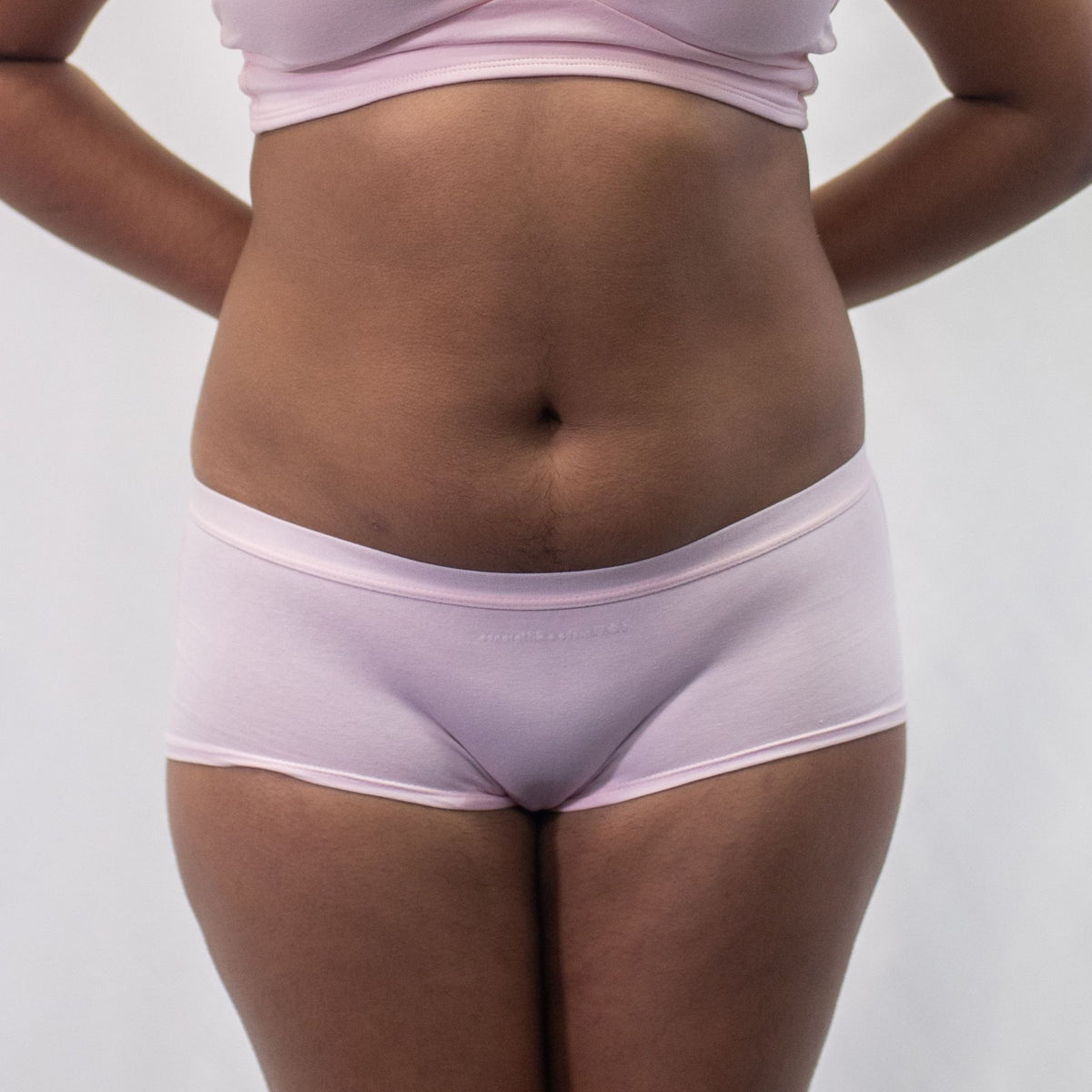 A photo of a model wearing the YOU Underwear boyshorts in pink. They are standing and facing the camera with their hands held behind their back. The photo is zoomed in so we can see from the model's stomach to their upper-thigh. The background is white.