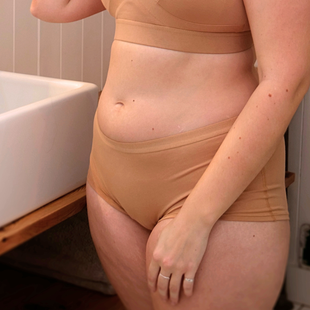 A person stands next to a sink, wearing comfortable, nude-colored boy shorts made from organic cotton. Their right hand rests on their hip while their left hand, adorned with a ring, rests near their thigh. The background includes part of a wooden counter and white wall paneling.