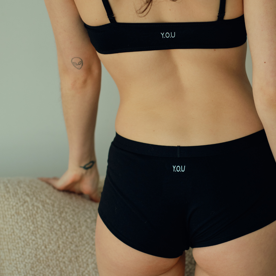 A person wearing a black bra and matching black boy shorts with the letters "Y.O.U" on them is leaning on a beige textured surface. The person has a tattoo of an alien head on their left arm and a tattoo of a ring on their left middle finger.