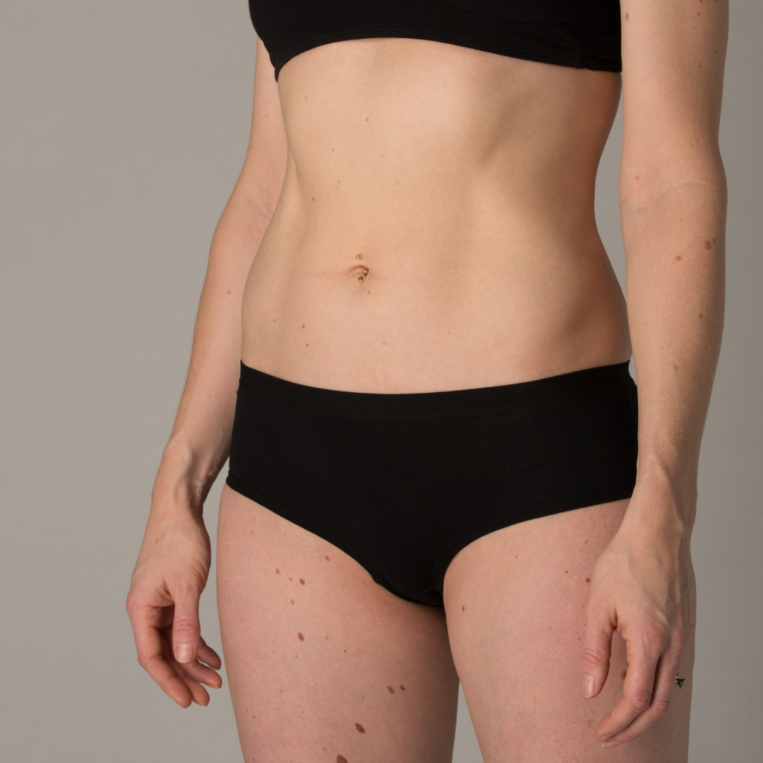 A person wearing black boy shorts and a black bralette is shown from the neck down. The individual has fair skin, a pierced navel, and visible moles on their torso and arms. The background is plain and grey.