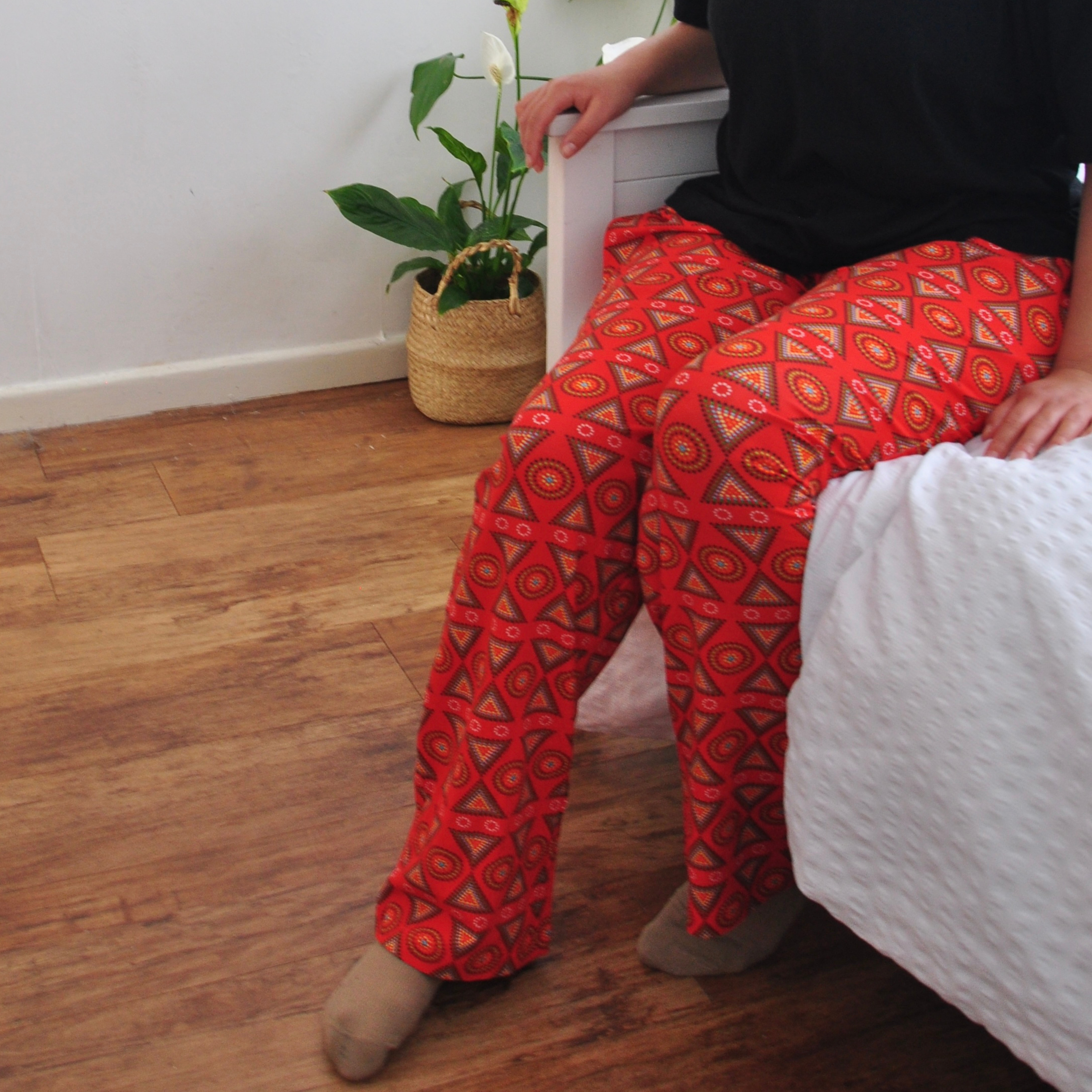 A person wearing vibrant, patterned red pyjama pants and a black shirt sits on the edge of a white bed with a textured blanket. In the corner, there is a woven basket holding a potted plant, adding a touch of greenery to the room.